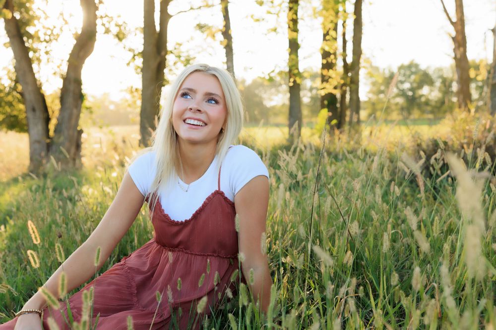 Someone smiles while sitting in a grassy field wearing a burgundy dress during sunset.