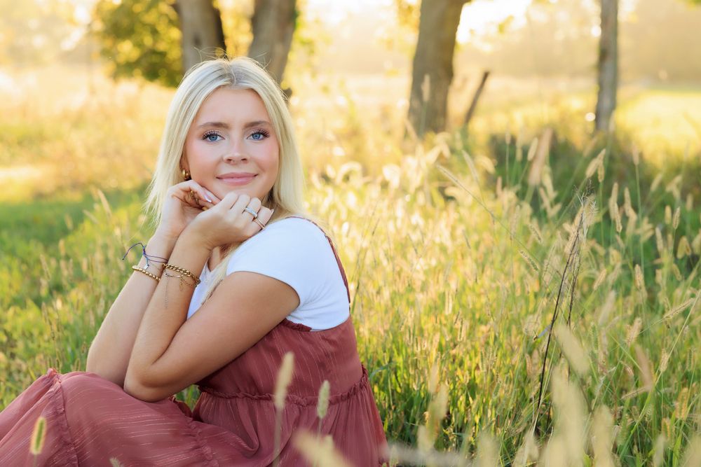 A person sits in a sunlit meadow wearing a white top and burgundy dress among tall grasses at golden hour.