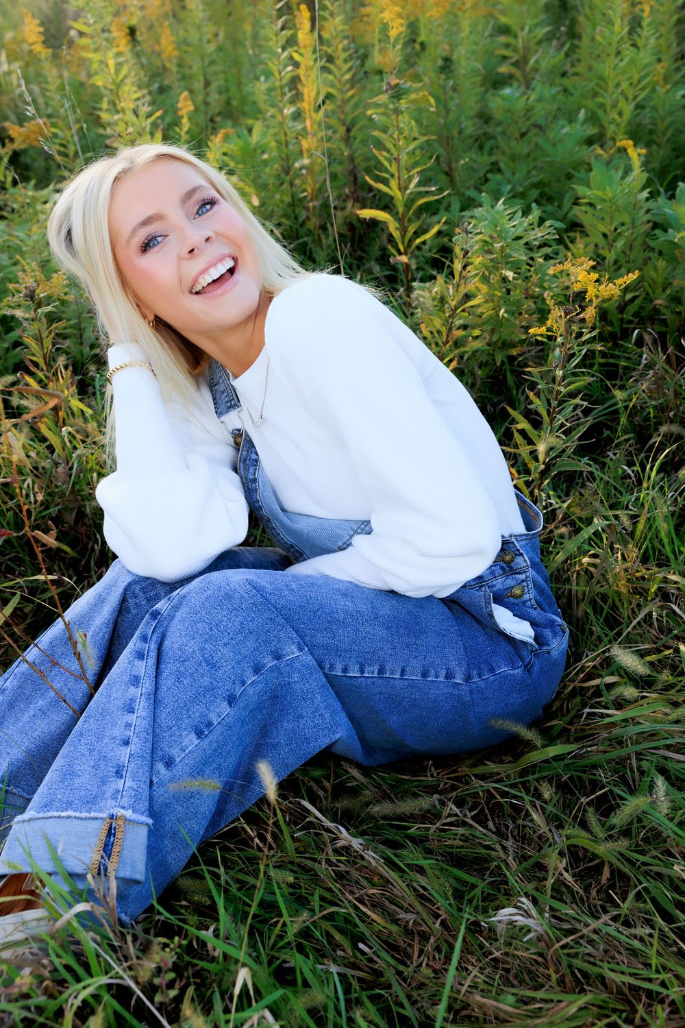 A joyful pose in a white sweater and denim overalls surrounded by tall wildflowers and greenery.