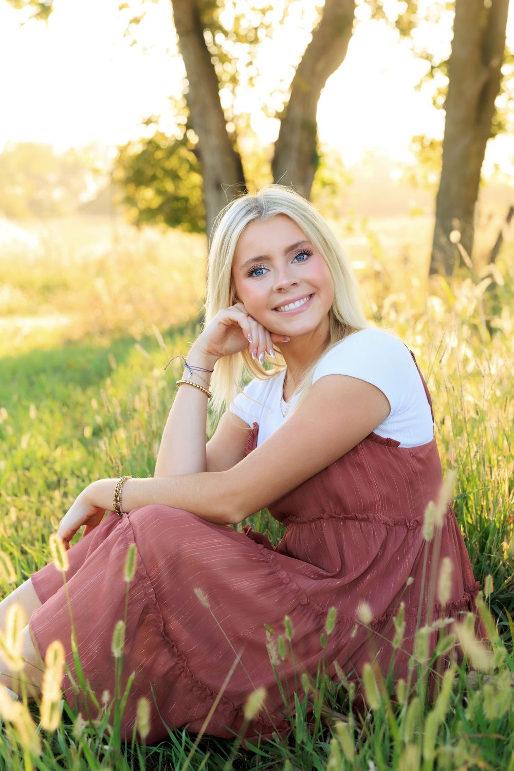Serene portrait in burgundy dress sitting among tall grass during golden hour.