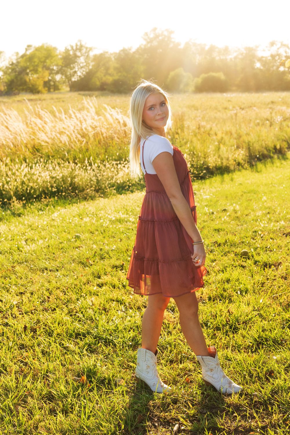 A full-length portrait shows someone in a burgundy dress and white sneakers standing in a sunny field.