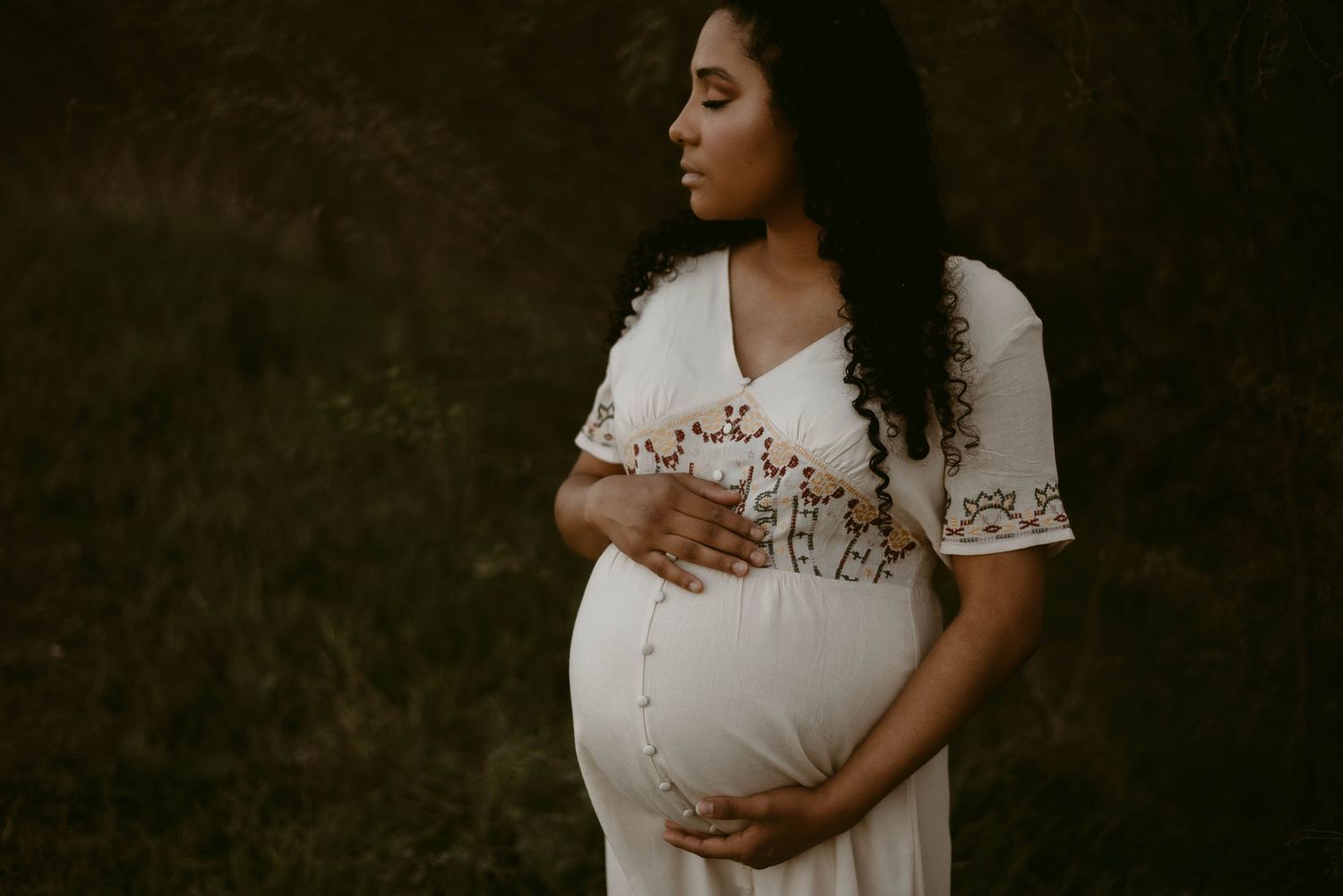 Dramatic side-lit maternity portrait featuring a white embroidered dress in a natural setting.