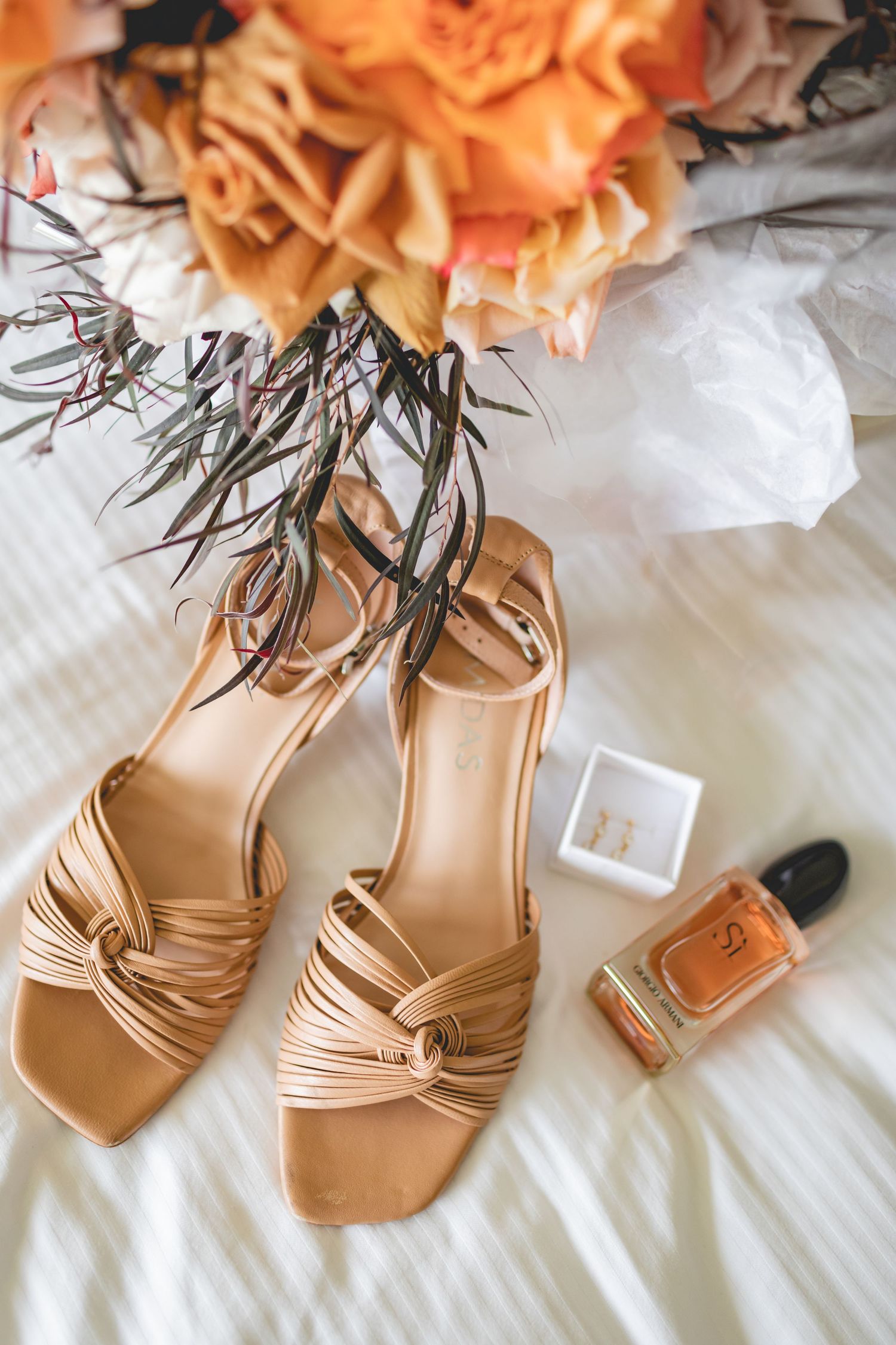 Neutral strappy sandals and nail polish arranged with peach floral bouquet on white bedding.