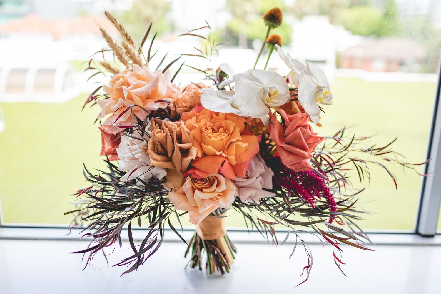 Elegant bridal bouquet with peach and coral roses mixed with dark wispy foliage against a window.