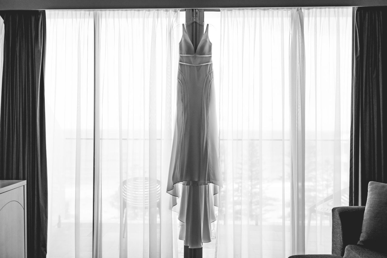 Wedding dress silhouette hanging in front of sunlit window with dark curtains.