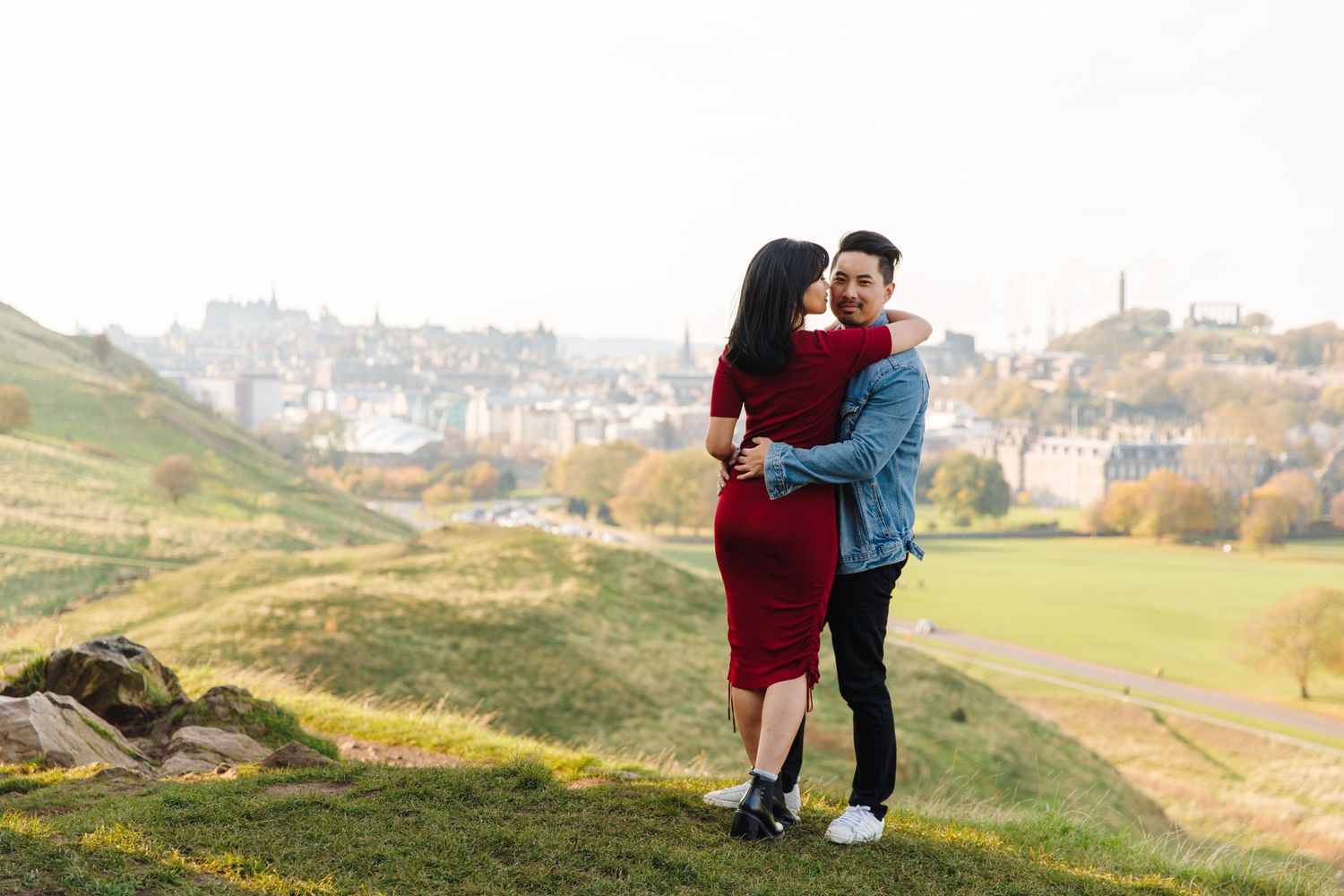 Couple sharing an embrace on a hilltop with Edinburgh's cityscape in the background.