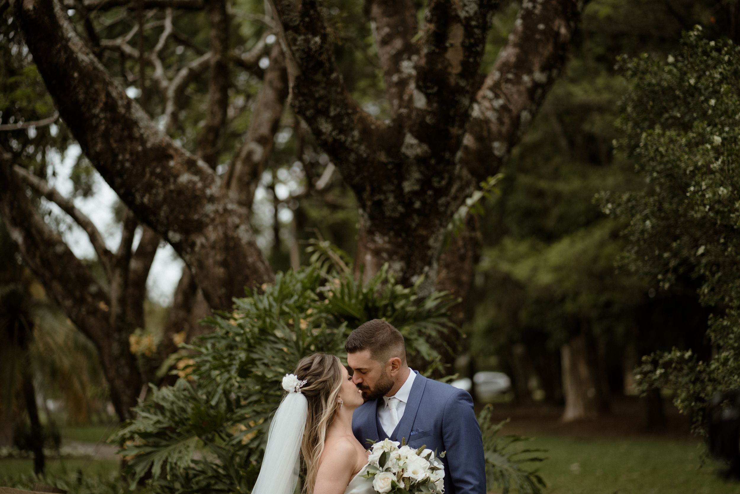 Romantic wedding portrait under towering trees in a lush garden setting.
