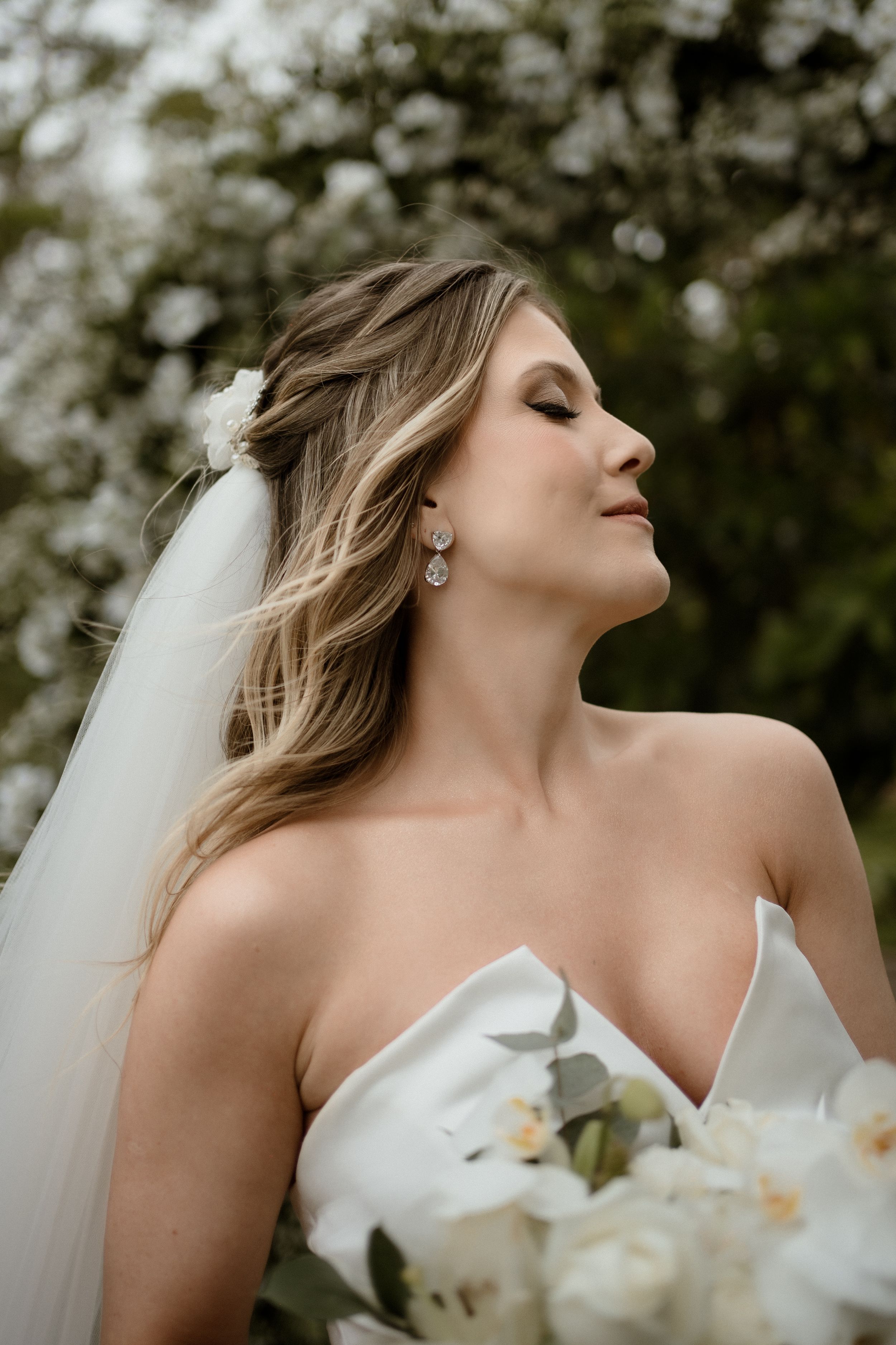 Elegant bridal portrait with flowing veil and white bouquet against natural outdoor backdrop.