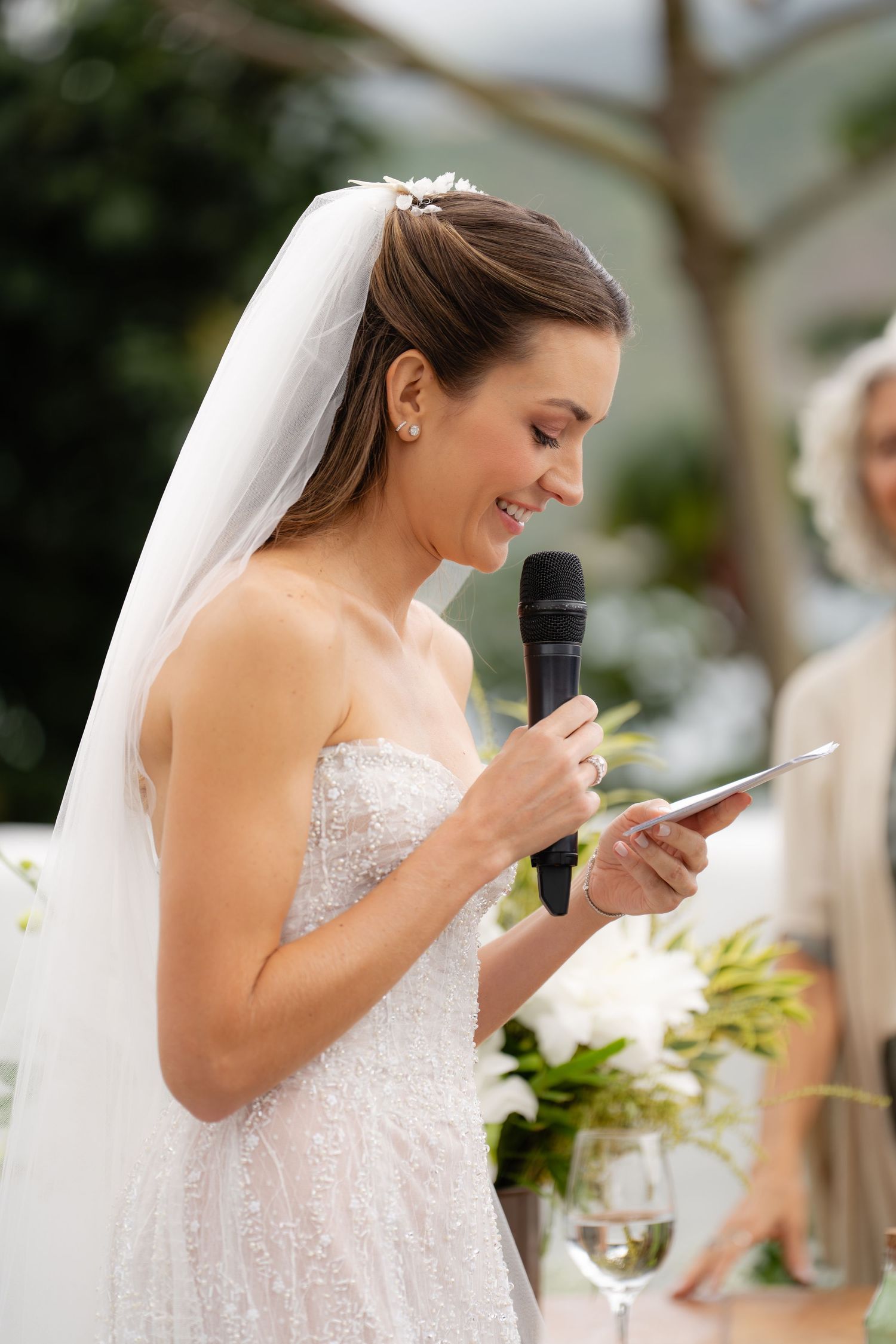 Bride in lace gown reads speech from notes during outdoor wedding ceremony.