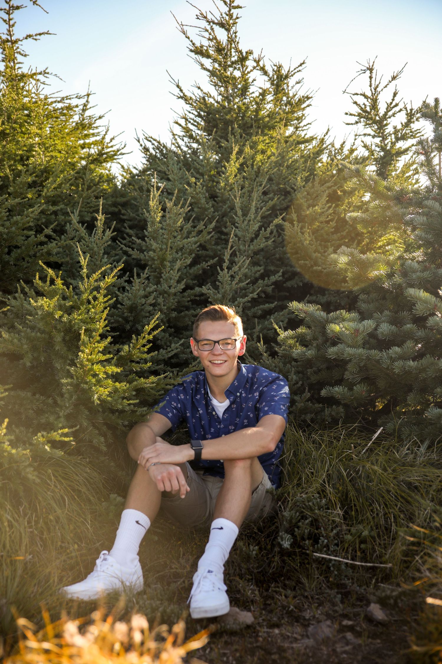 Person sitting among evergreen trees during golden hour wearing a blue patterned shirt and white socks.