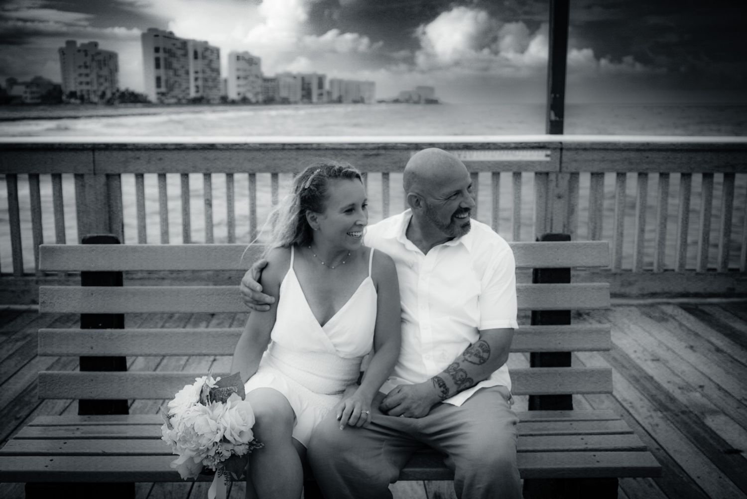 Black and white photo of a bride and groom sitting on a wooden bench near the beach.