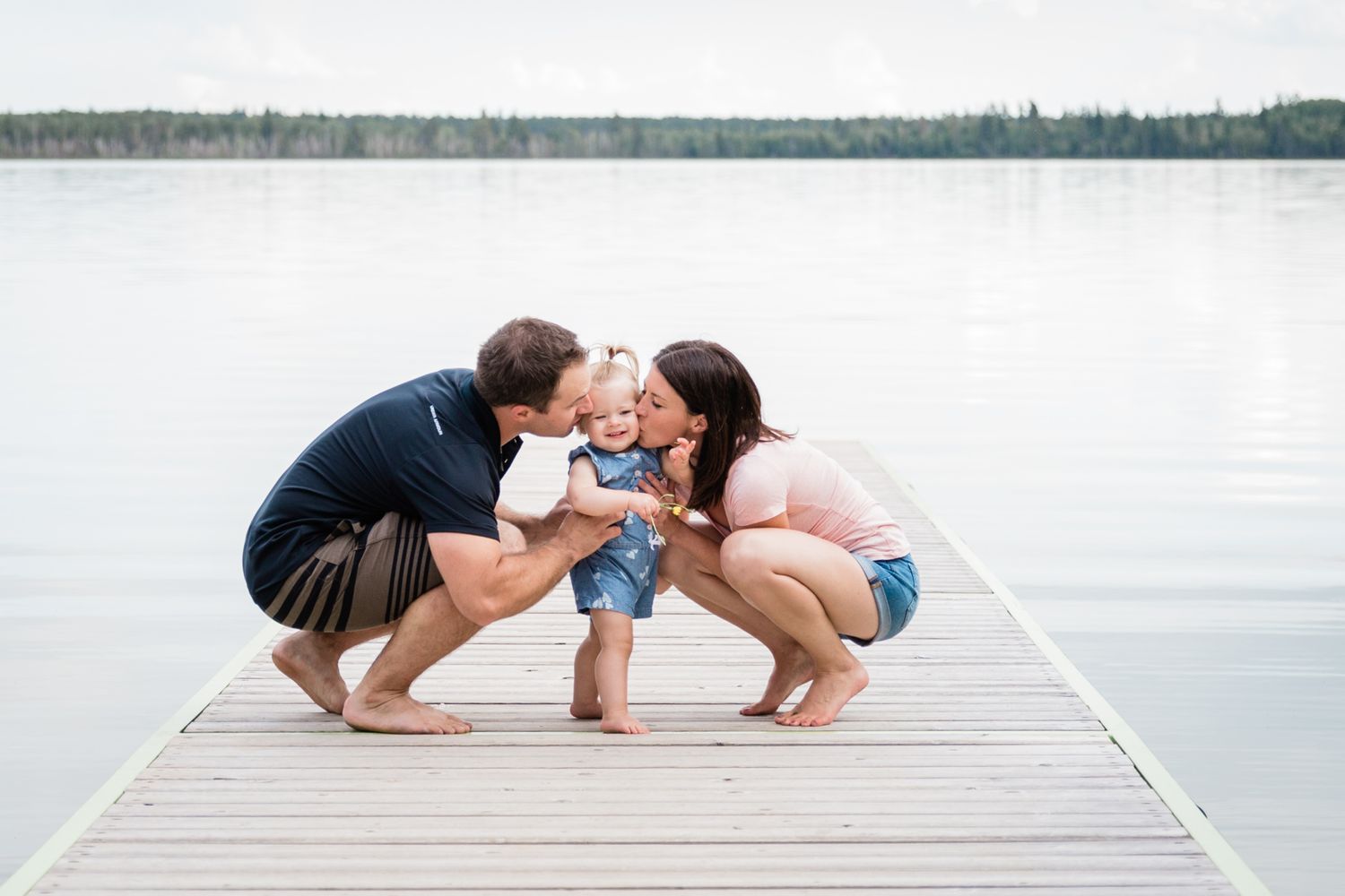 Happy parents kiss their baby on a wooden dock by a serene lake on a cloudy day.