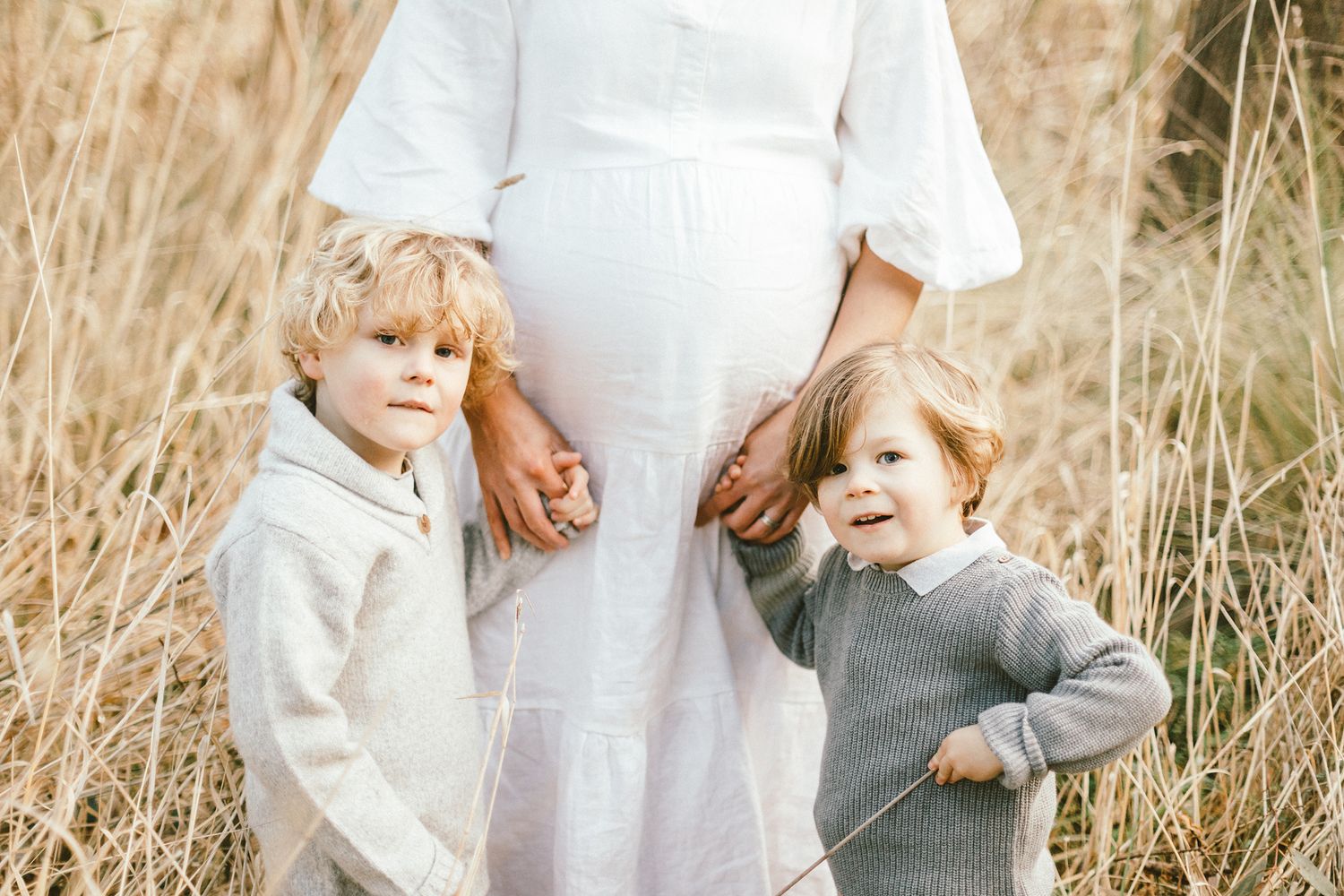 Family portrait with expectant mother in white dress standing among tall grass.