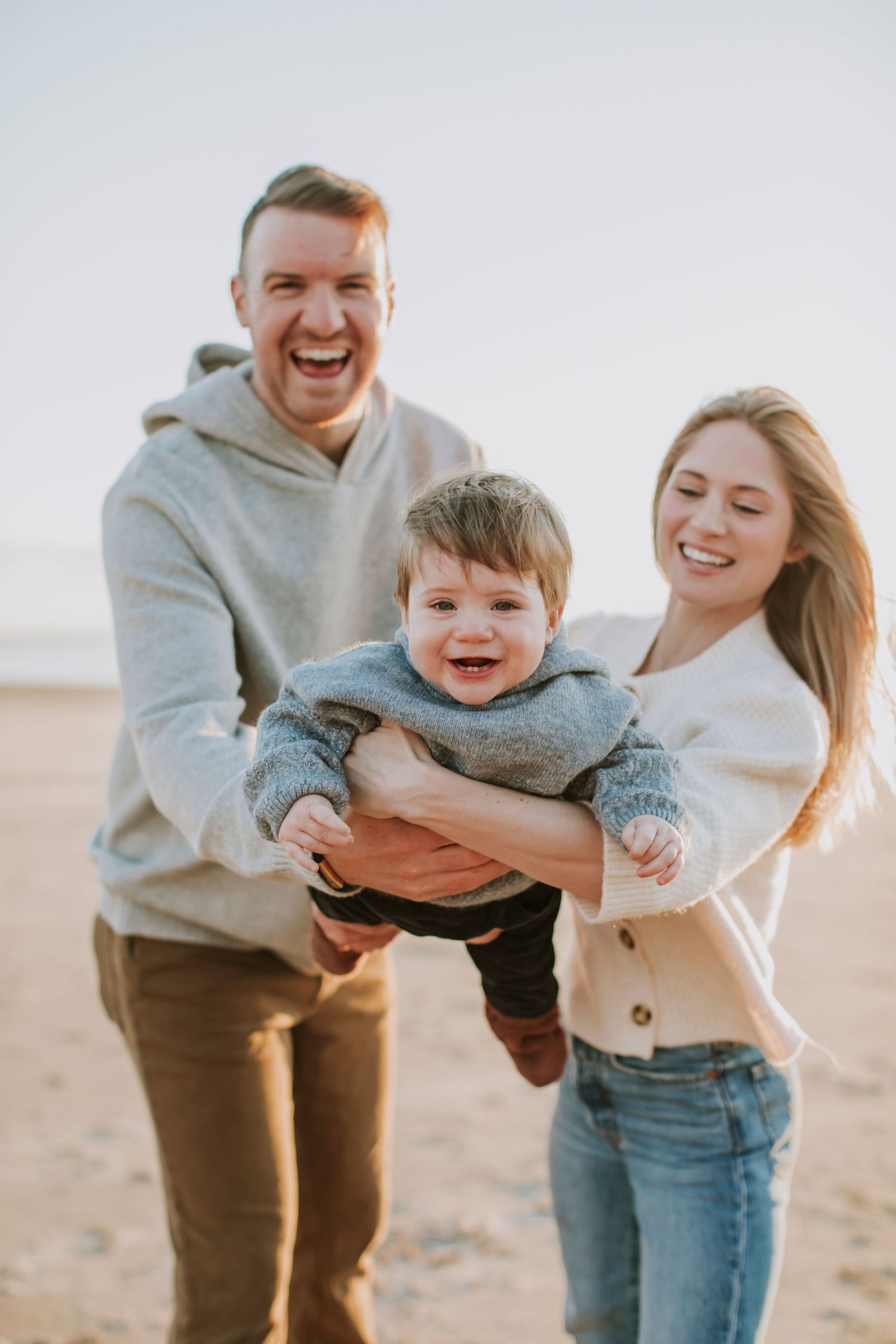 A mom and dad laughing while swinging their smiling baby boy in front of them at the beach