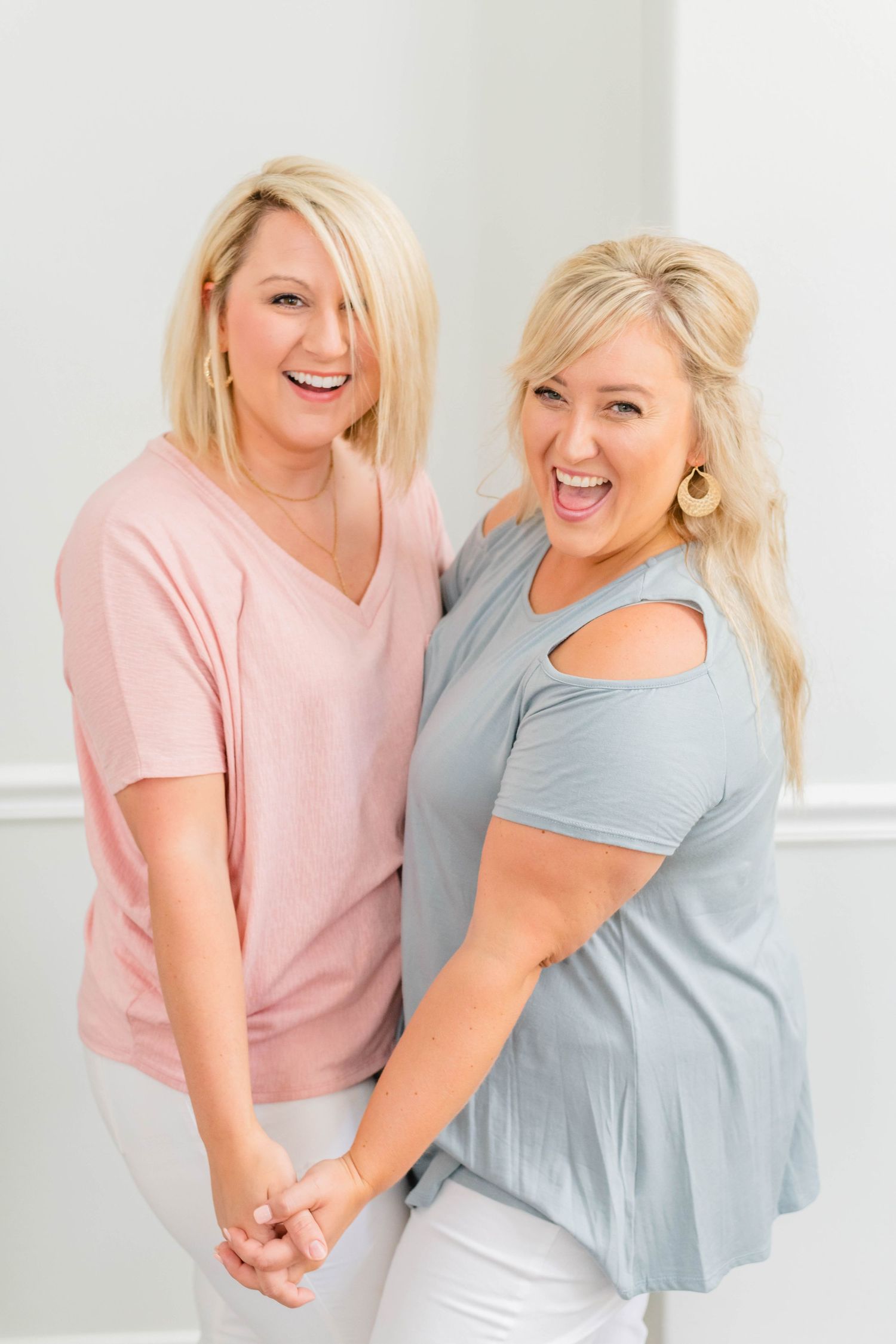 Two smiling women, one wearing a pink shirt and the other wearing a gray shirt, stand together and hold hands. Katie Gibbons Photography