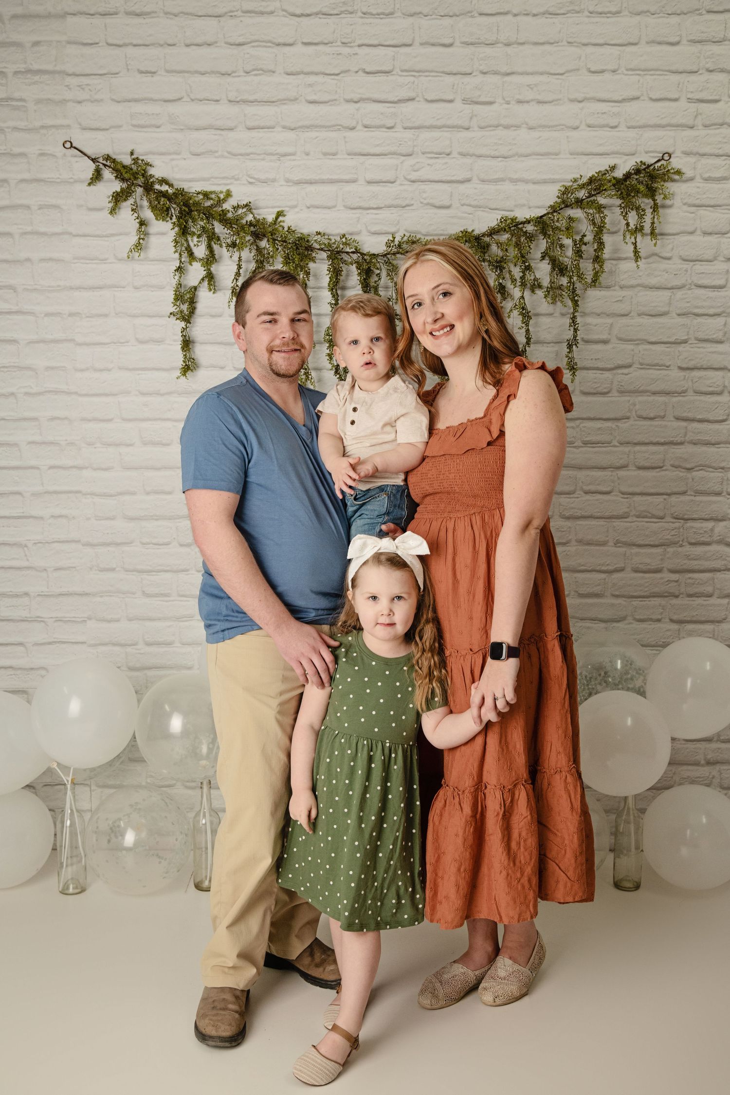 Family portrait against white brick wall with evergreen garland and white balloons.