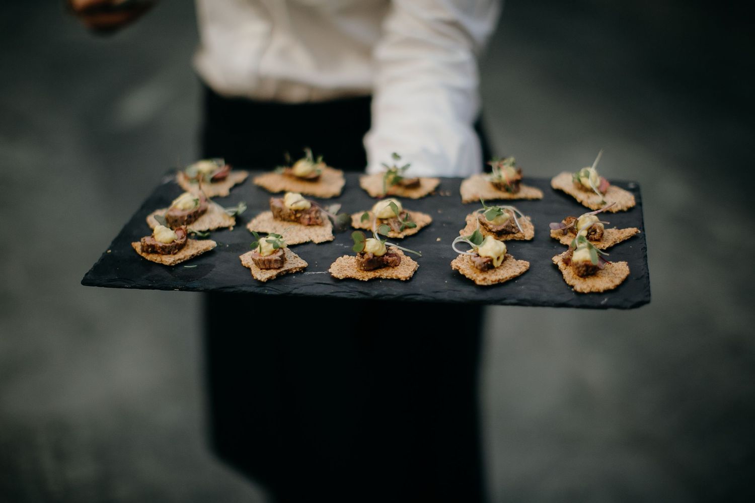 waiter hold a tray of brown rice crackers with meat and sauce on a charcoal serving board