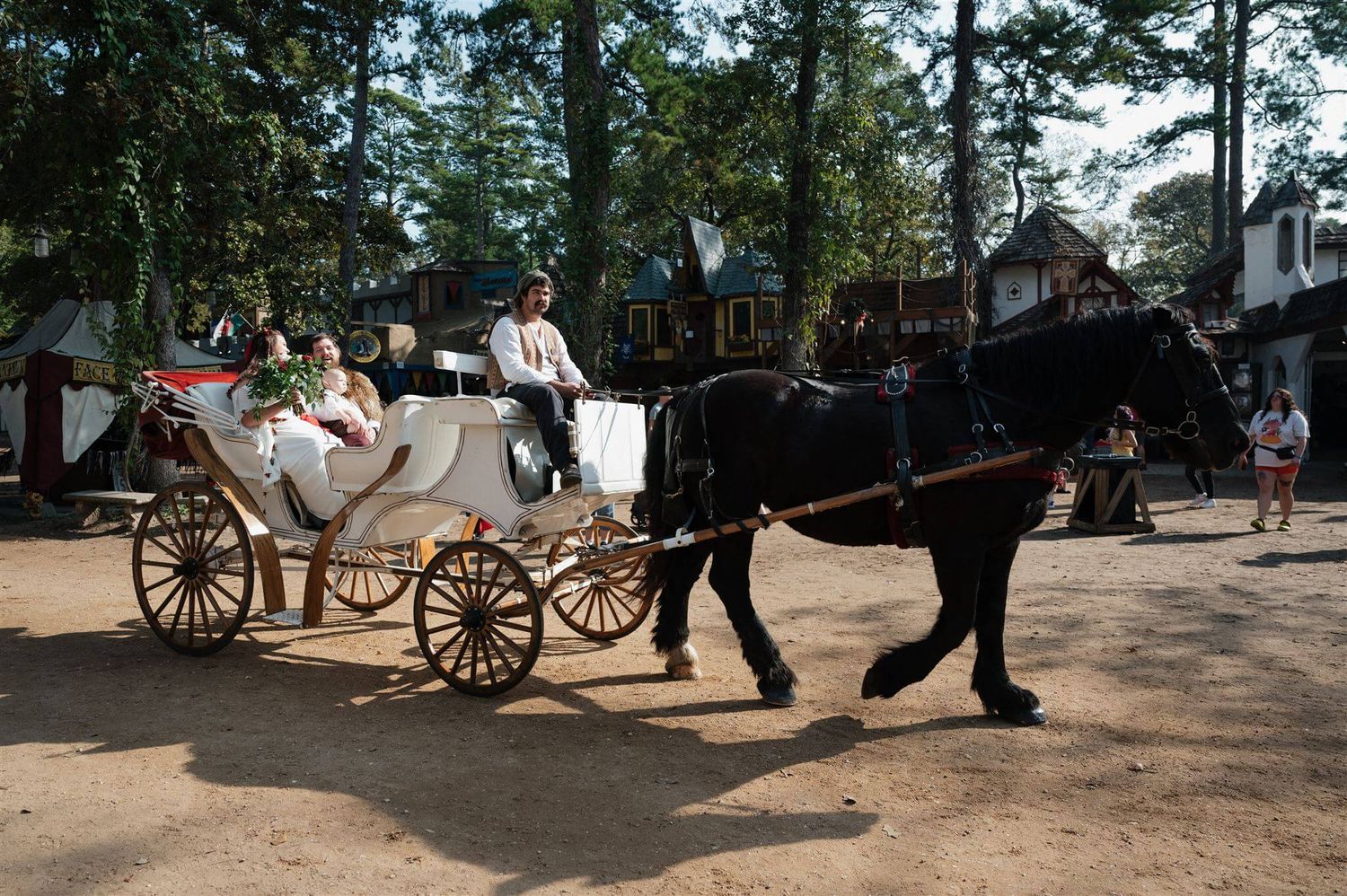 A white horse-drawn carriage carries passengers at a renaissance faire.