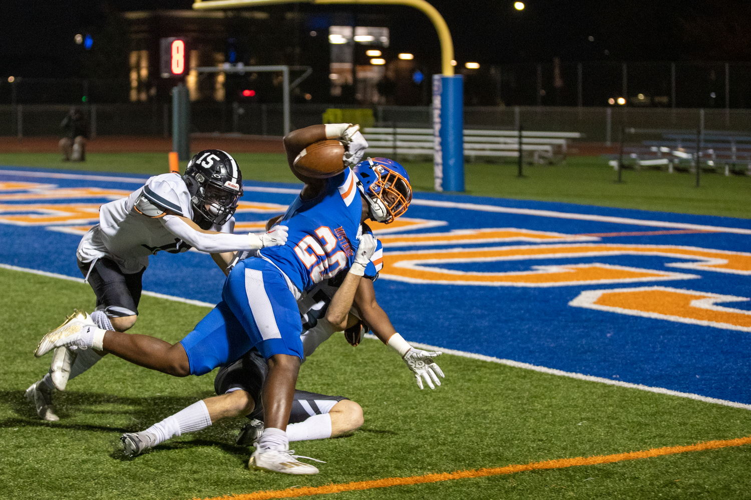 Football players competing for the ball on a brightly lit field with blue and orange markings.