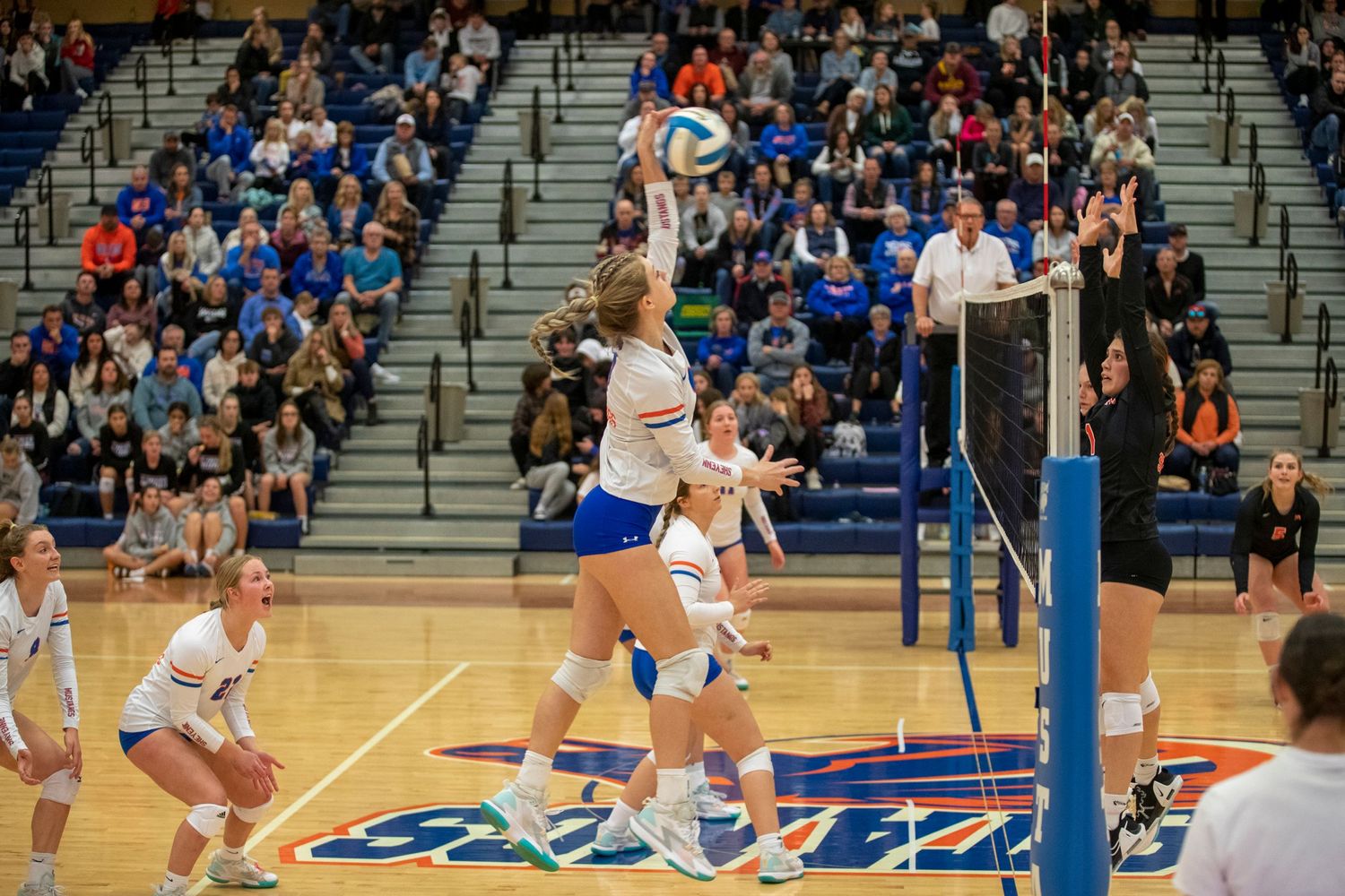 Indoor volleyball match with players jumping for the ball at the net.