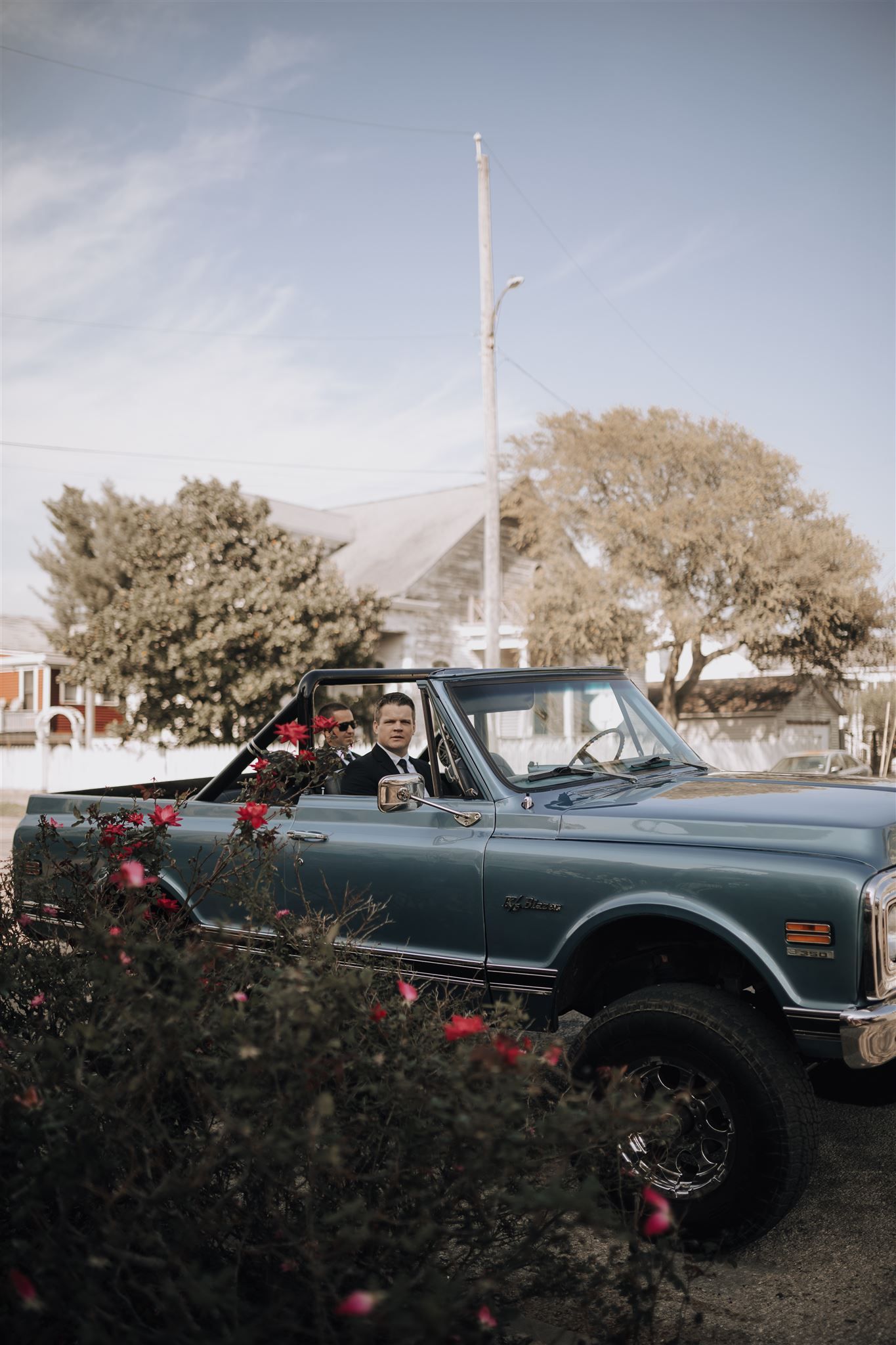 A vintage blue lifted truck with large tires parked near blooming red flowers.