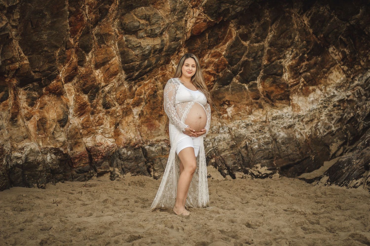 Beach maternity portrait against dramatic rocky cliffs at sunset.