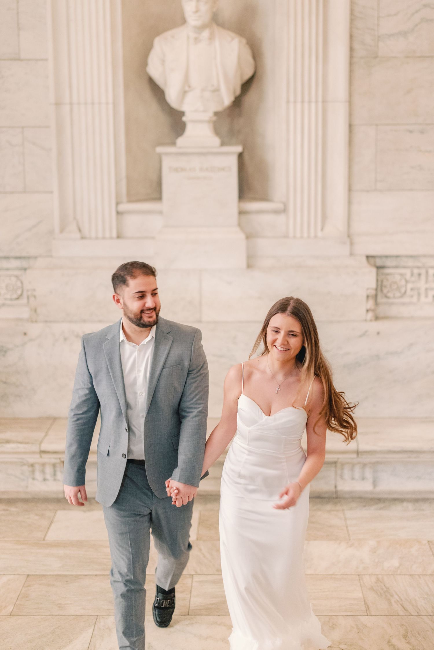 Smiling couple walking hand in hand past classical marble bust sculpture.