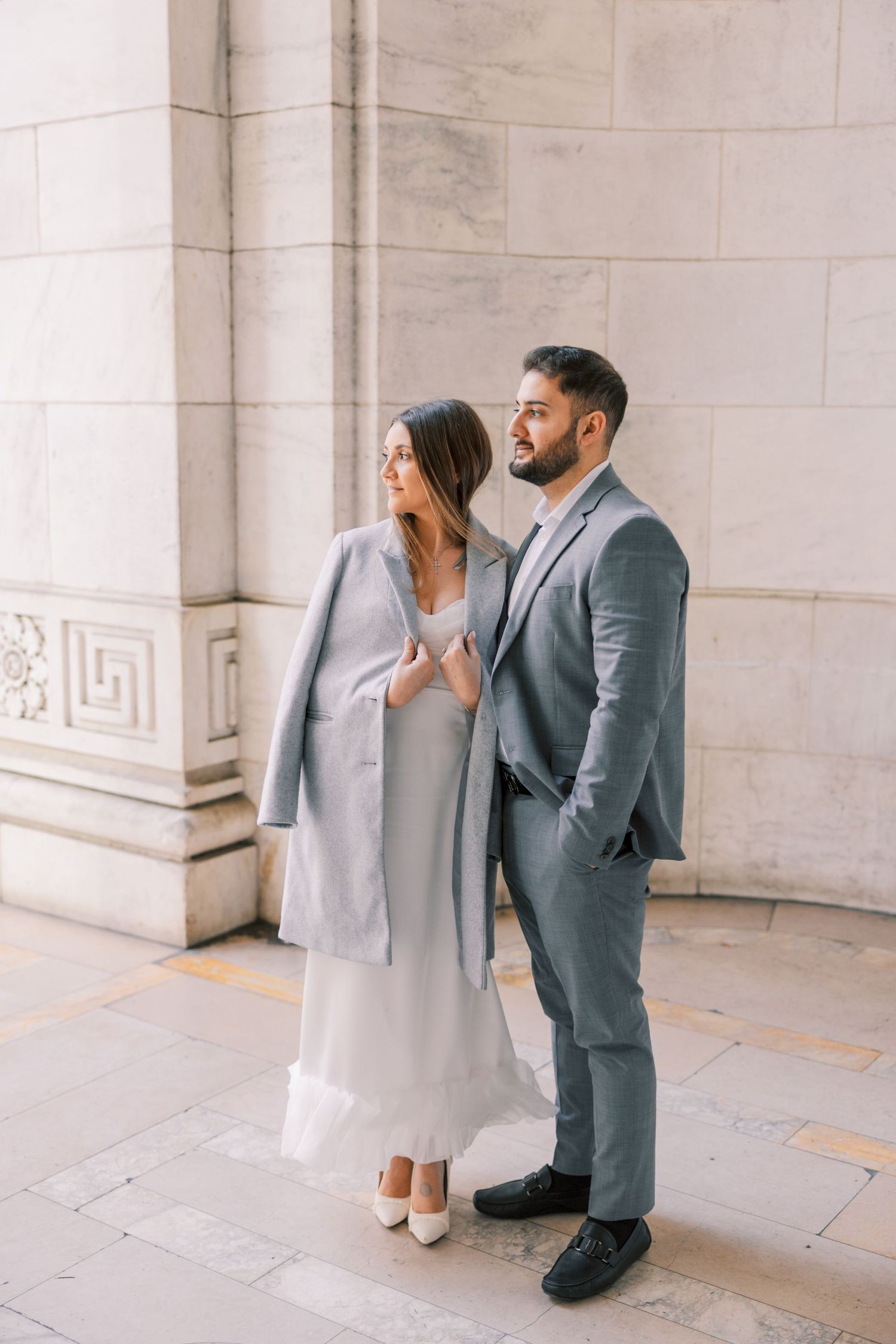 Elegant couple posing against marble wall, woman in white dress and gray coat, man in gray suit.