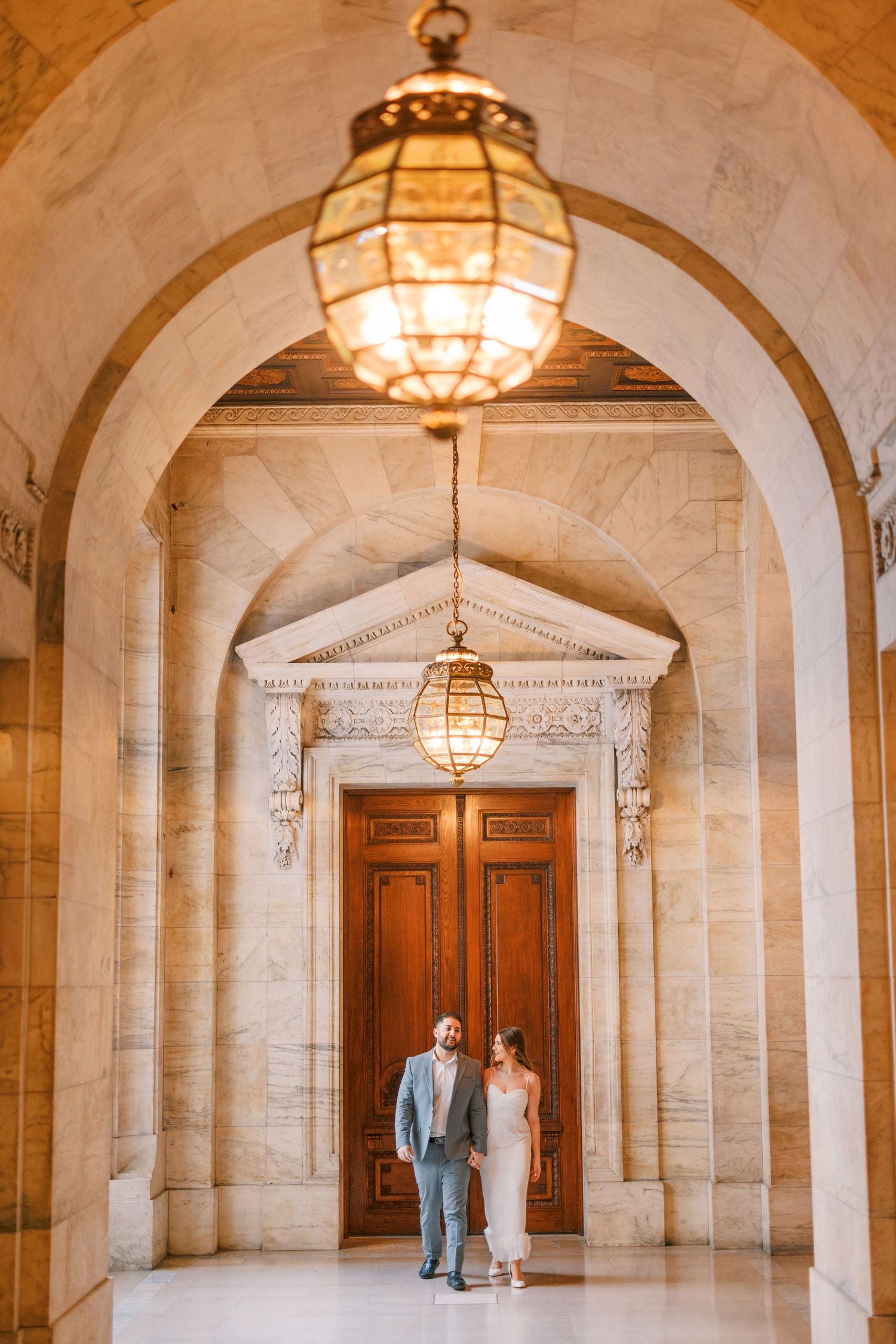 Couple standing beneath ornate chandelier in grand marble archway before elegant wooden doors.