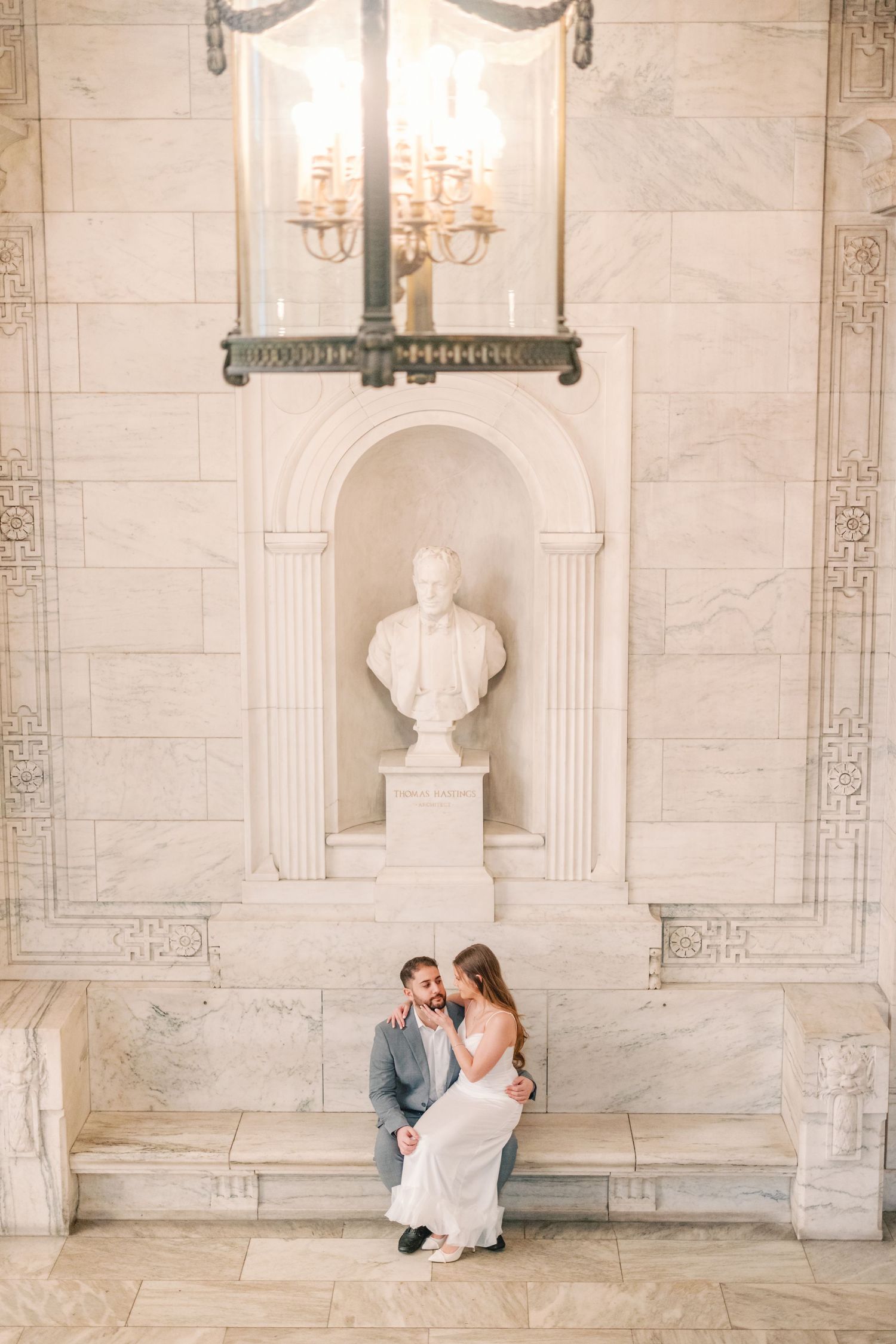 Newlyweds sitting together beneath marble bust sculpture in grand hall.