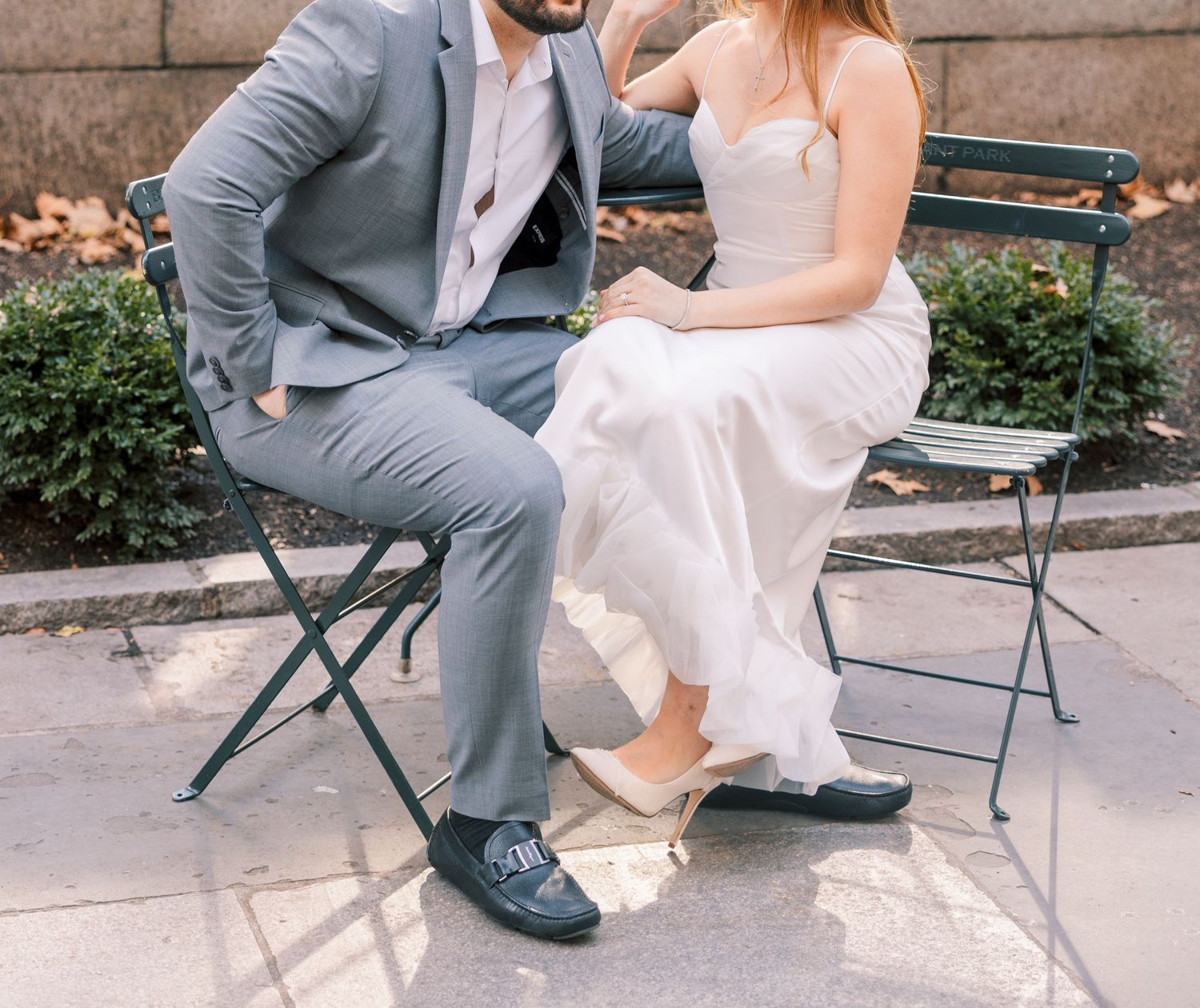 Couple seated on park bench, man in gray suit, woman in white wedding dress showing shoes.