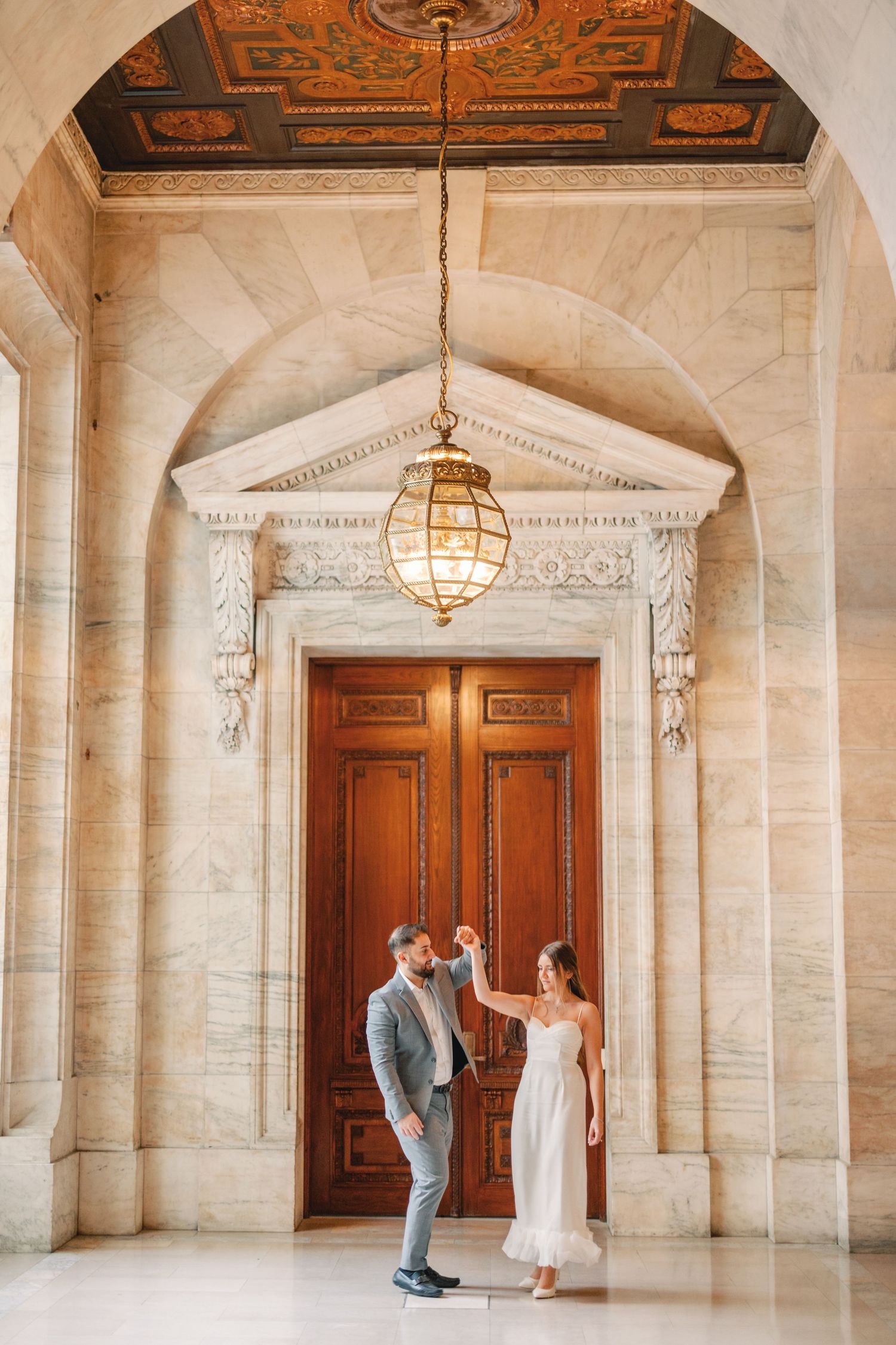 Groom spinning bride beneath vintage chandelier in elegant marble hallway with wooden doors.