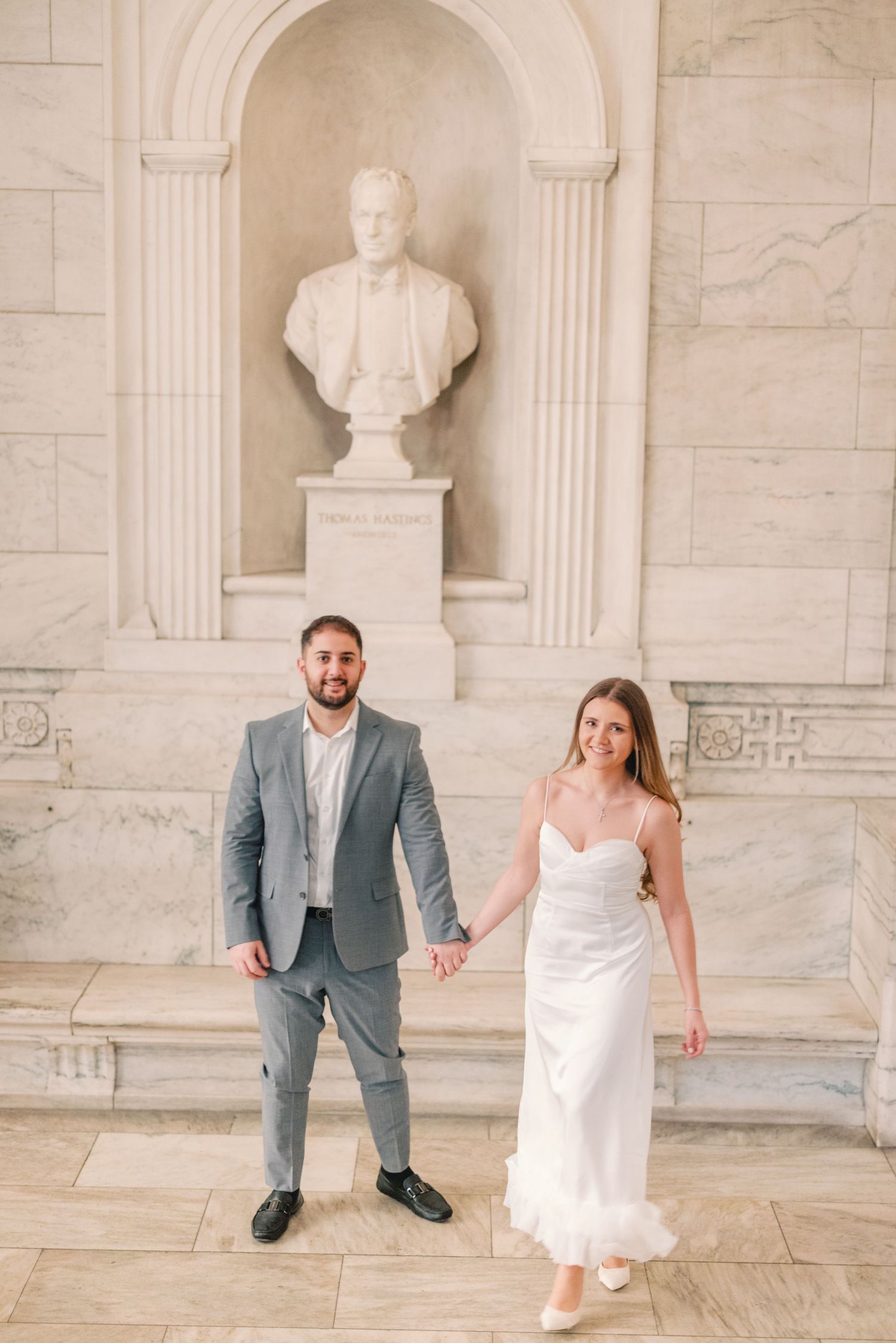 Bride and groom holding hands before marble bust in elegant venue.