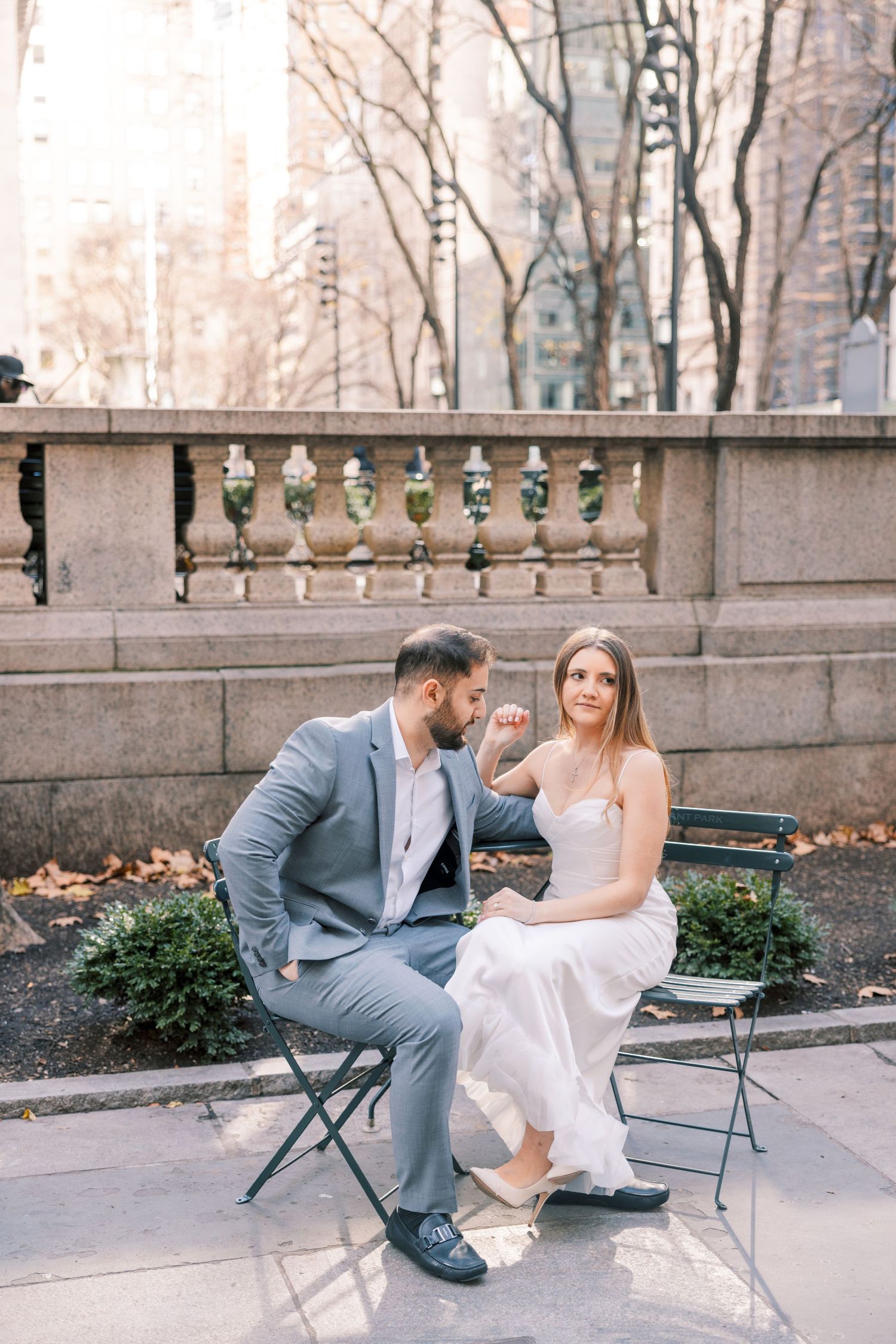 Groom kissing bride's hand while seated on garden bench with stone balustrade backdrop.