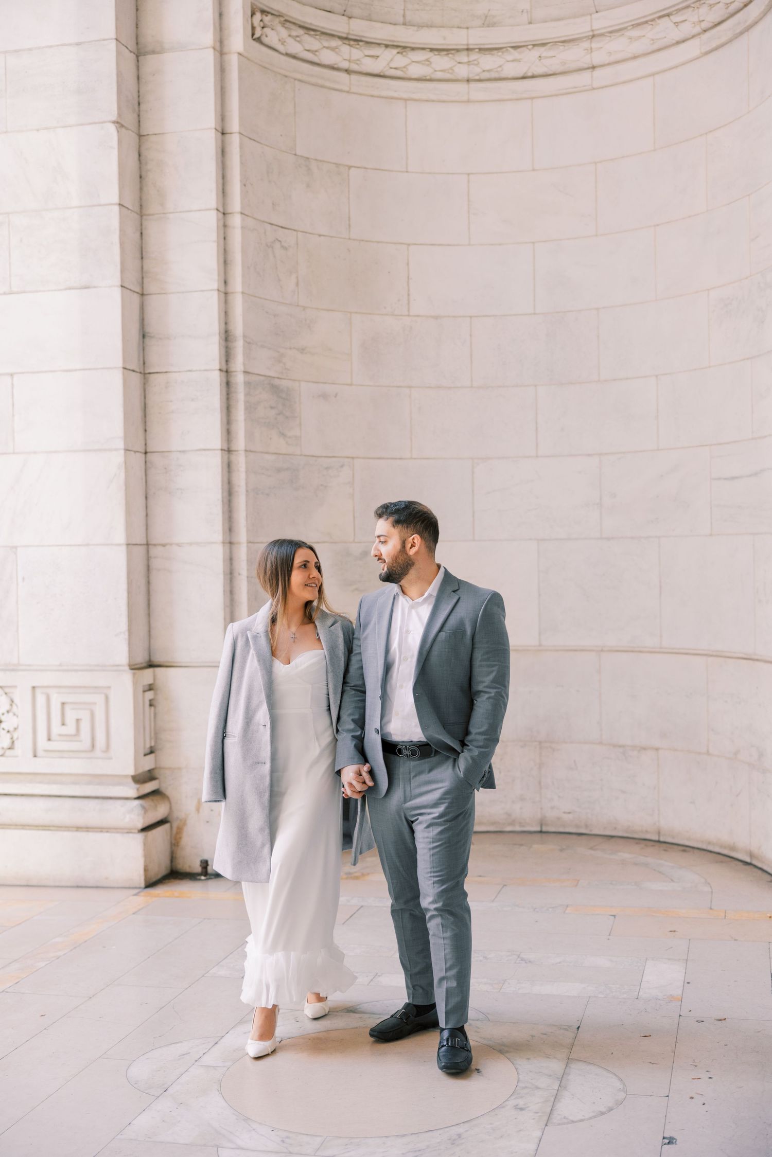 Stylish couple walking hand in hand near ornate marble columns in coordinating gray outfits.