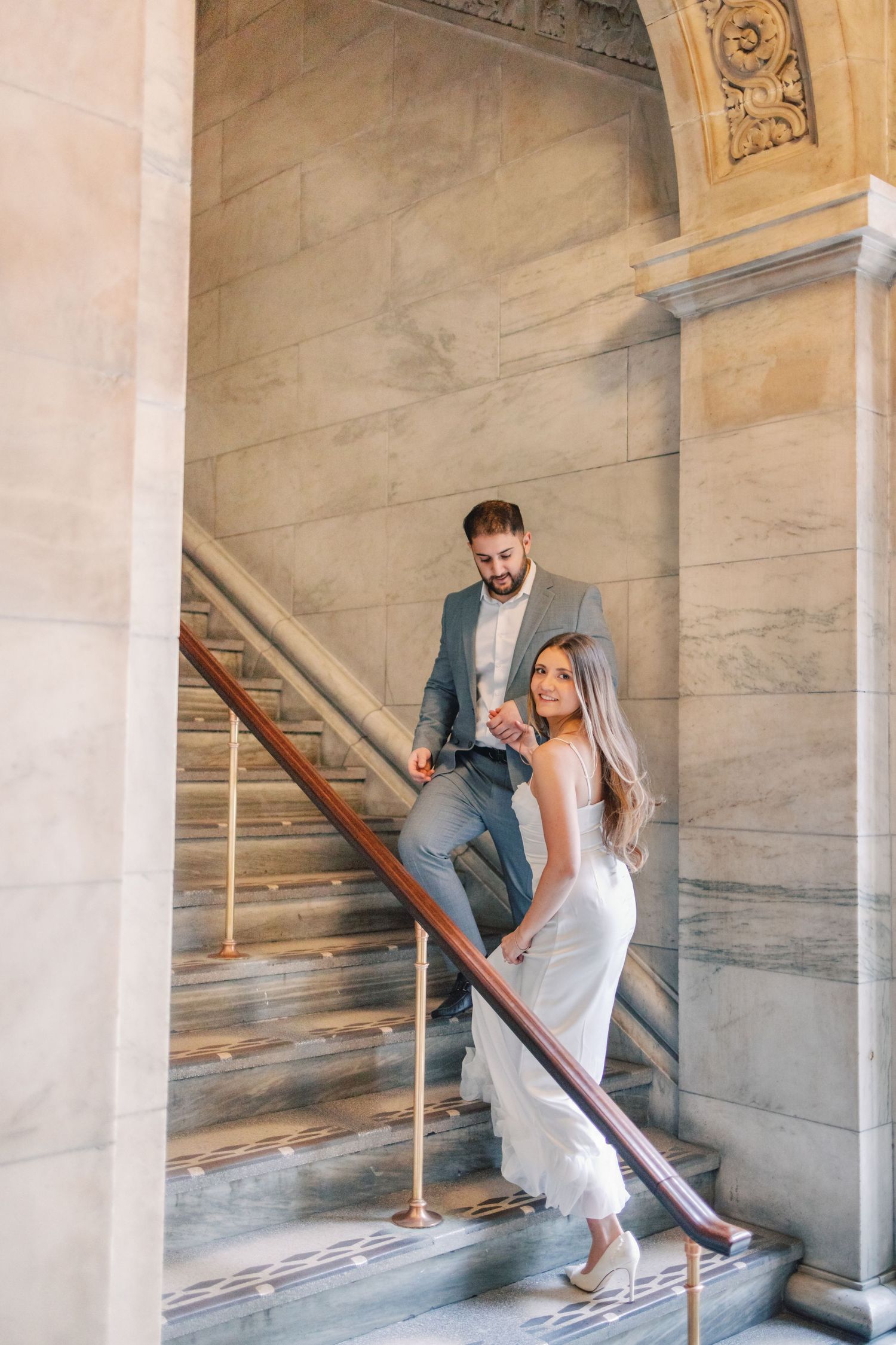 Bride and groom descending elegant marble staircase in historic building.