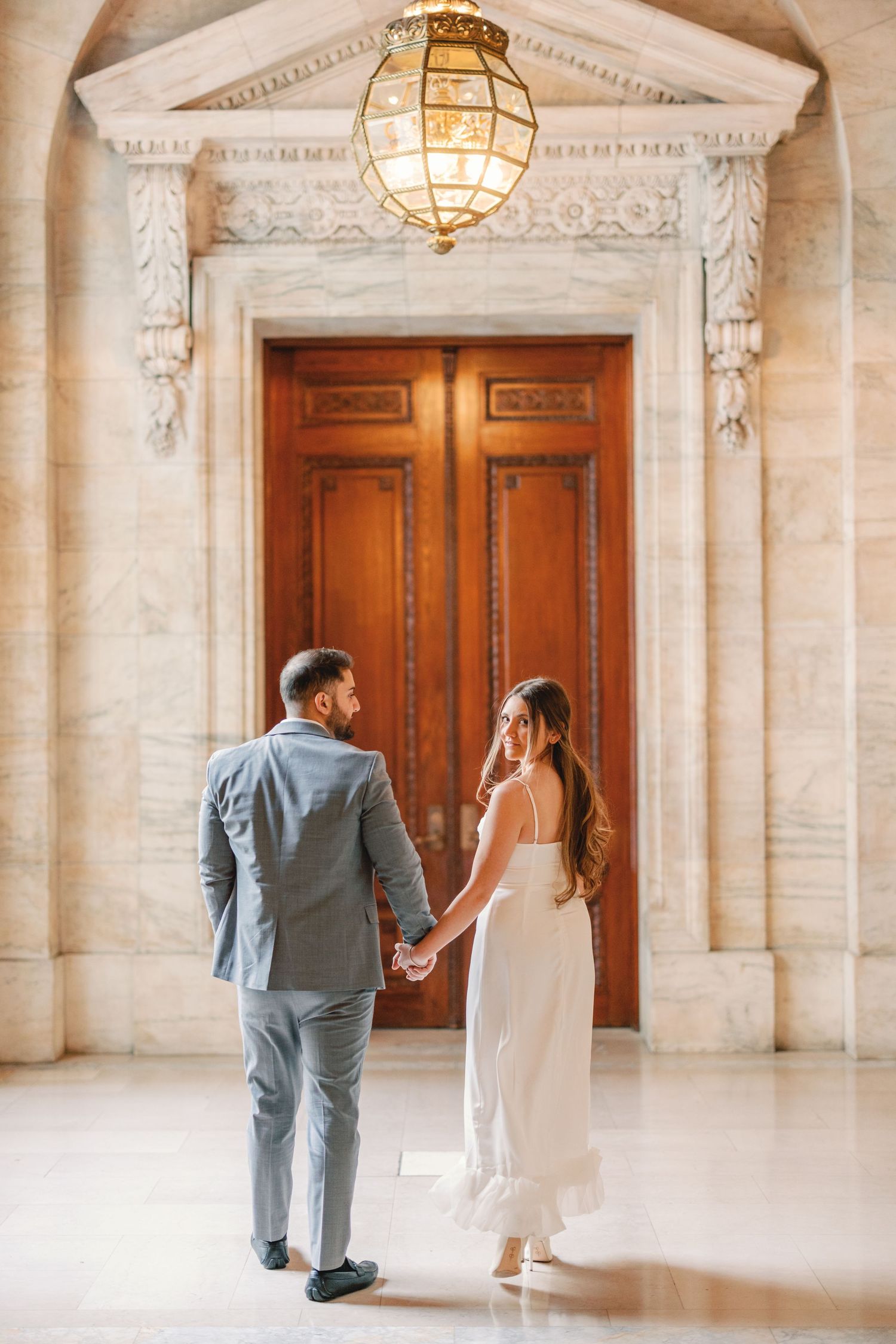 Couple holding hands facing ornate wooden doors under glowing lantern in marble corridor.