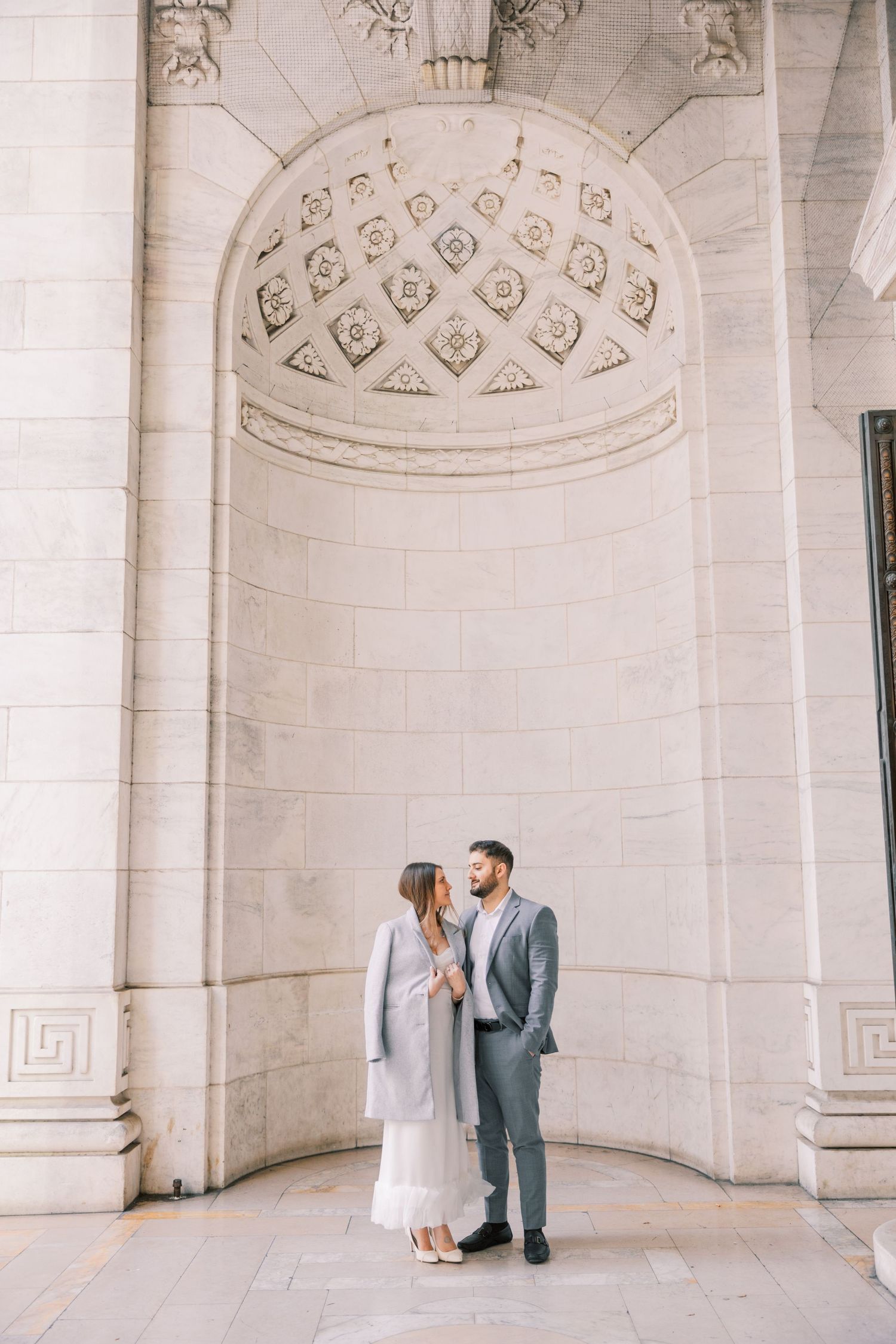 Engaged couple shares tender moment beneath ornate stone archway of a grand neoclassical building exterior.