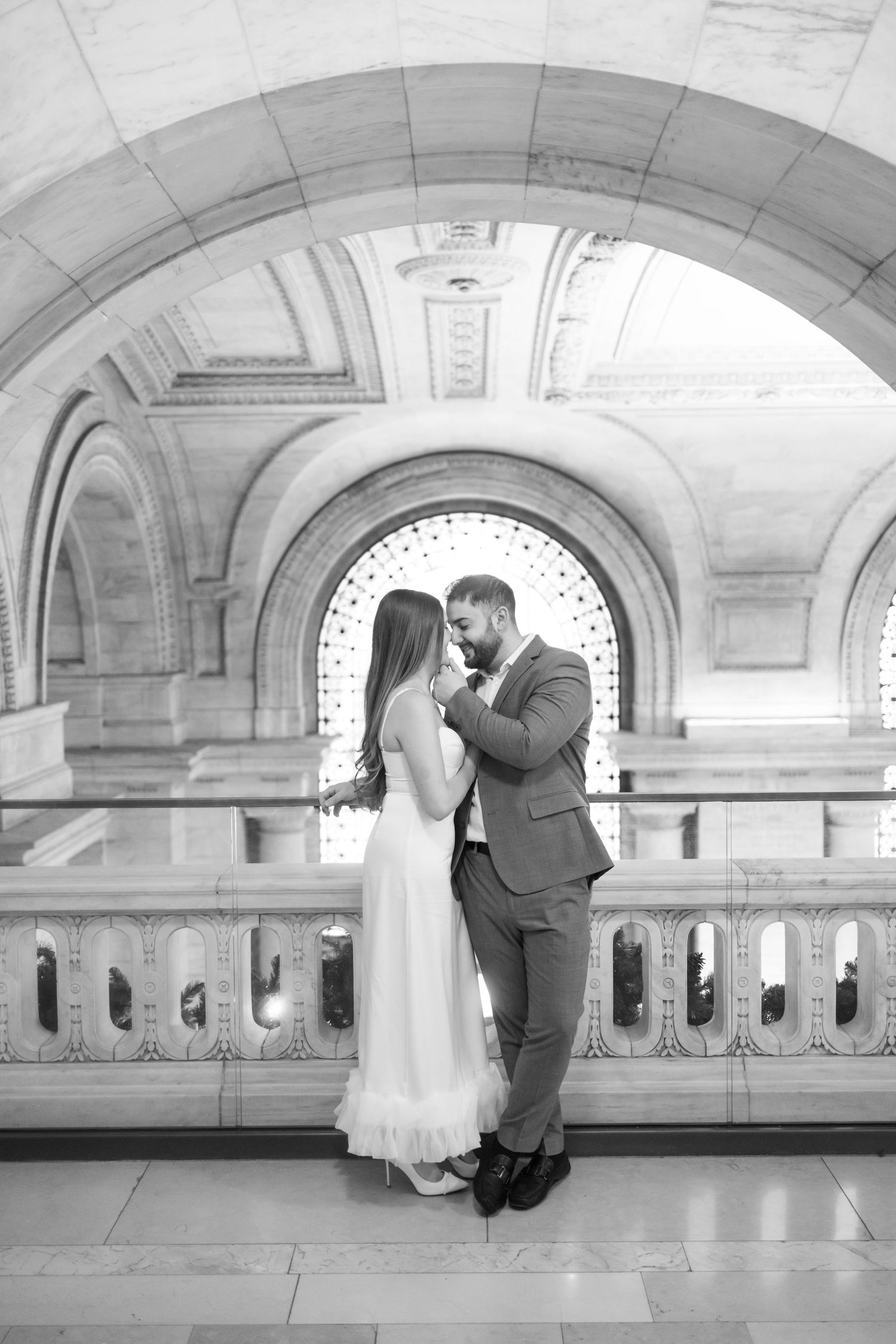 Black and white romantic portrait of couple embracing under grand arched window in marble hall.