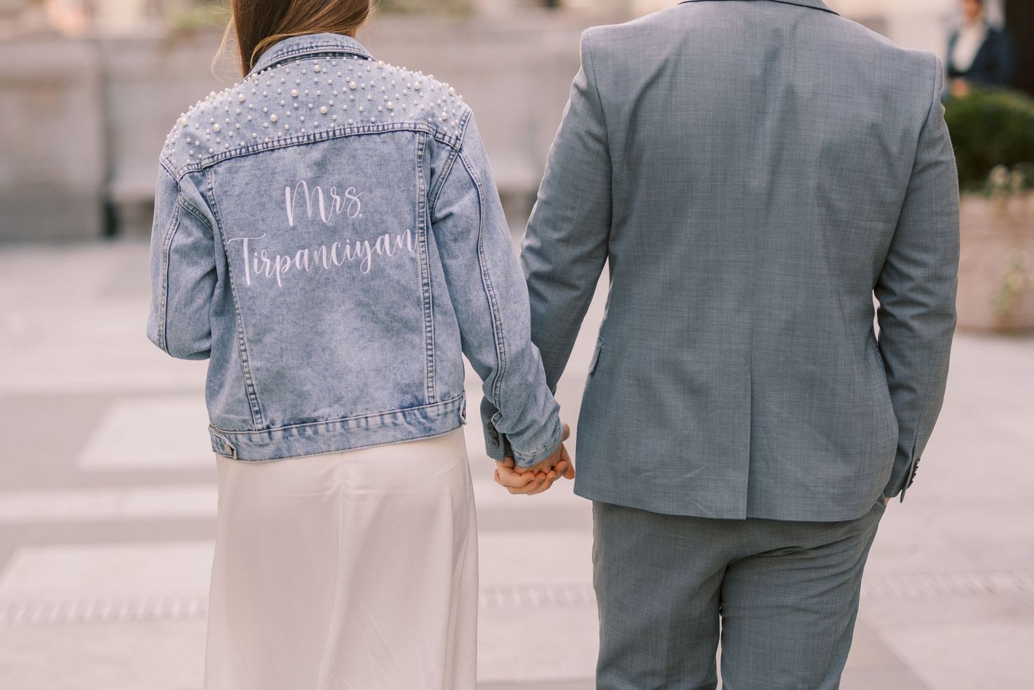Close-up of bride wearing custom Mrs. denim jacket holding grooms hand.