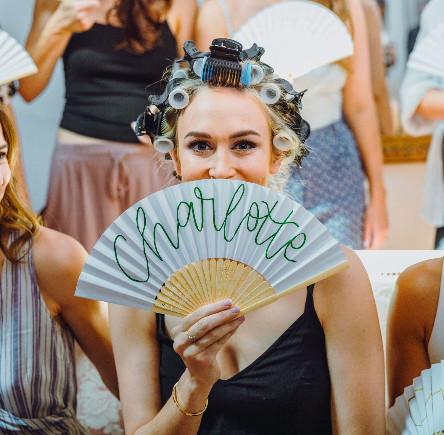 Wedding guest with rollers in hair holds up decorative Charlotte-themed hand fan at celebration.