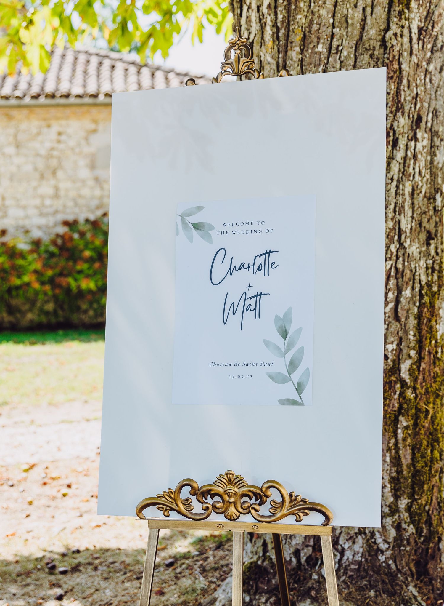 Elegant wedding welcome sign with floral design displayed on gold stand against tree trunk.