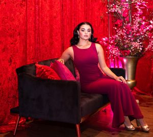 Woman in burgundy evening gown poses elegantly on black couch against red backdrop with floral arrangement.