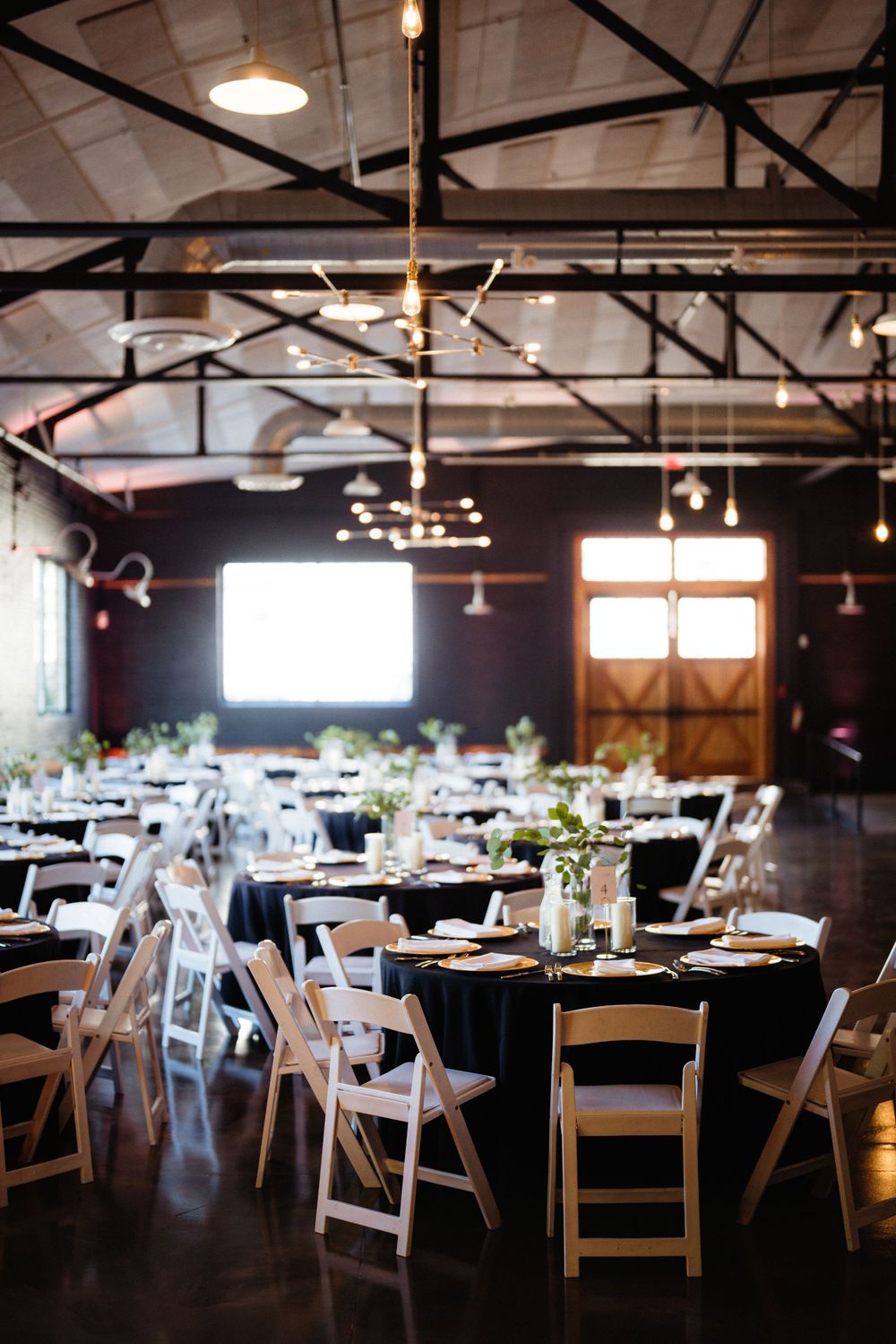 An elegant wedding reception venue with round tables, white chairs and industrial ceiling lighting.