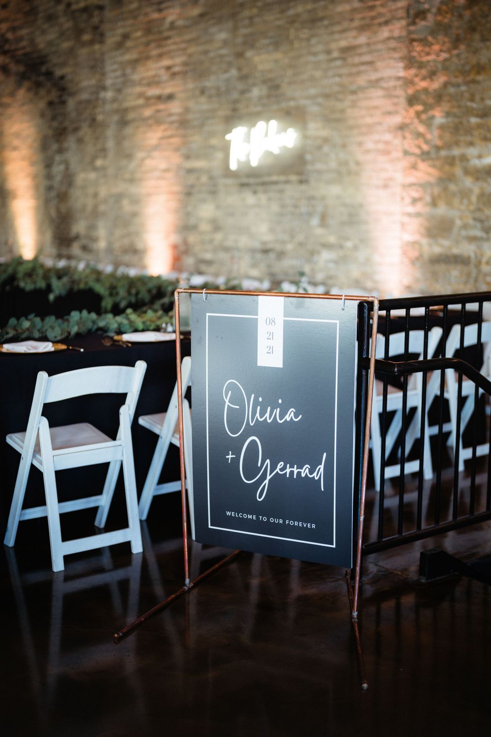 Wedding reception venue sign with white folding chairs and illuminated brick wall background.