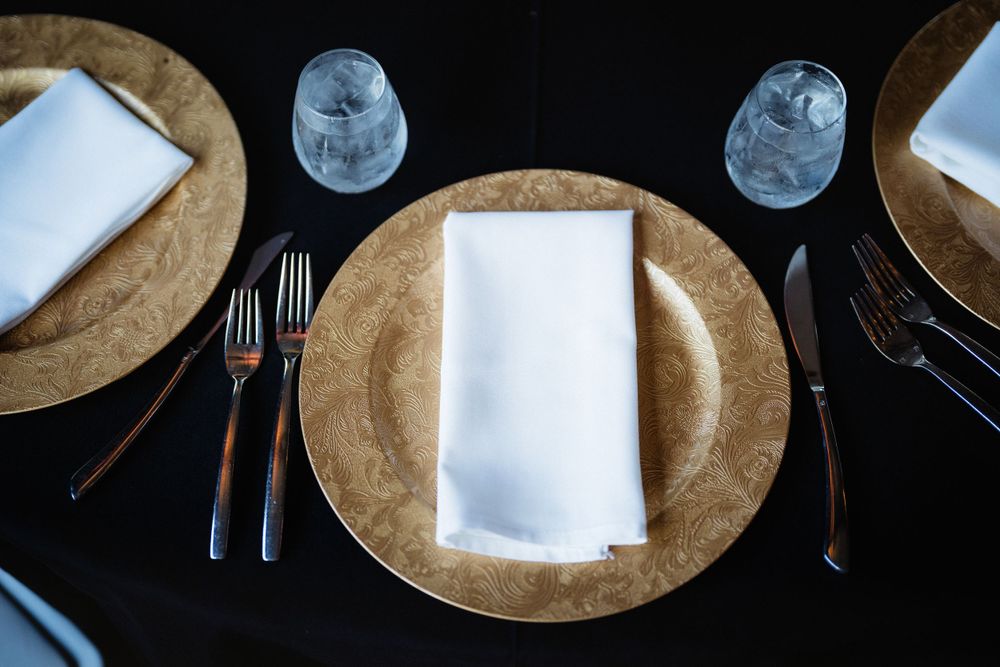 Place setting with gold charger plate, white napkin, and silverware on black tablecloth.