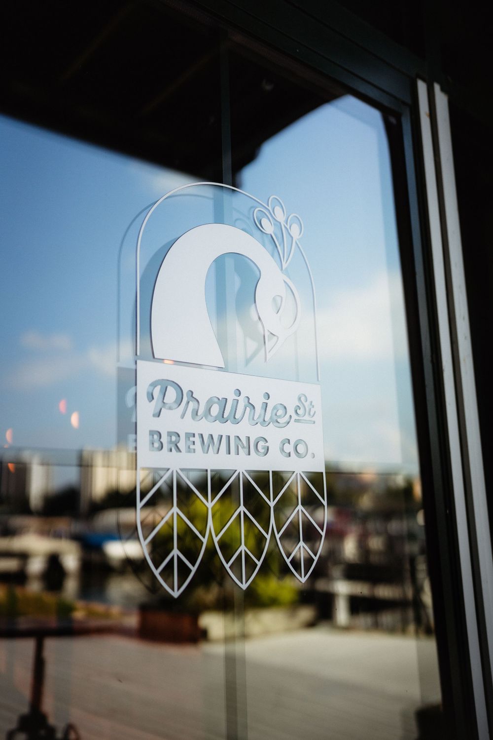 Prairie Brewing Company logo etched on glass window with blue sky background.