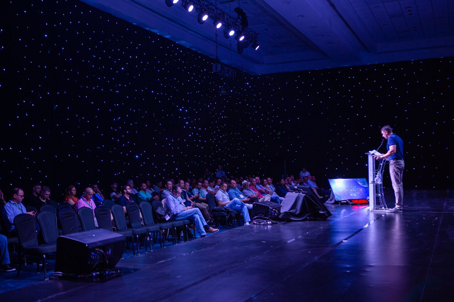 Speaker presenting on stage to an audience in a dark auditorium with blue lighting.
