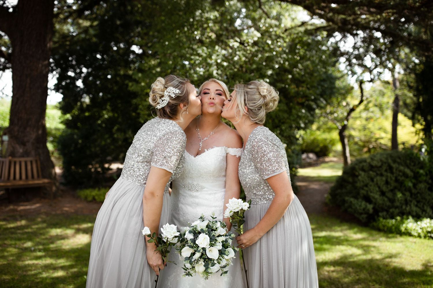 Bridesmaids in silver dresses share sweet moment with bride in garden setting.