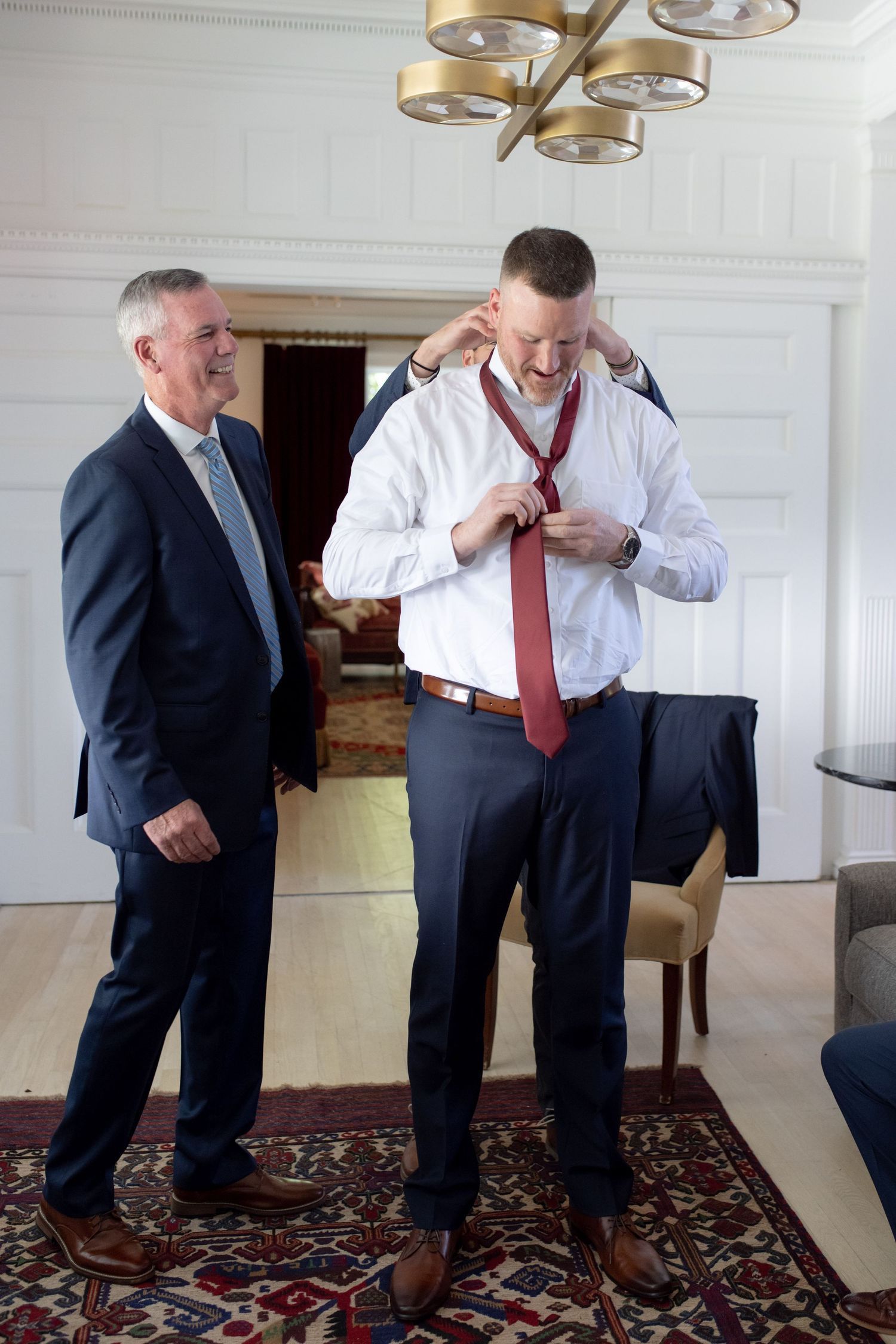 Two men in formal attire prepare for an event in an elegant room with ornate lighting.